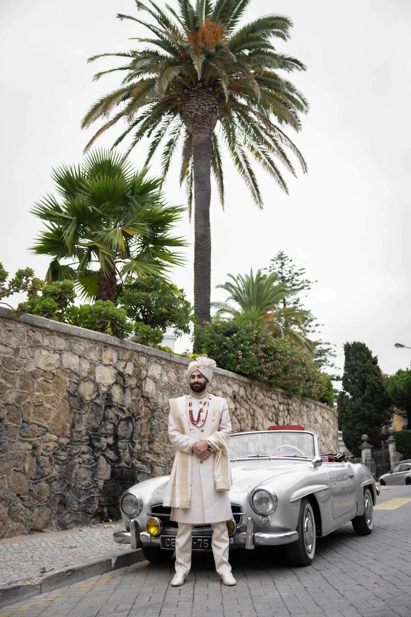 A man dressed in traditional Indian attire standing beside a vintage silver car on a cobblestone street, with a stone wall, palm trees, and lush greenery in the background.