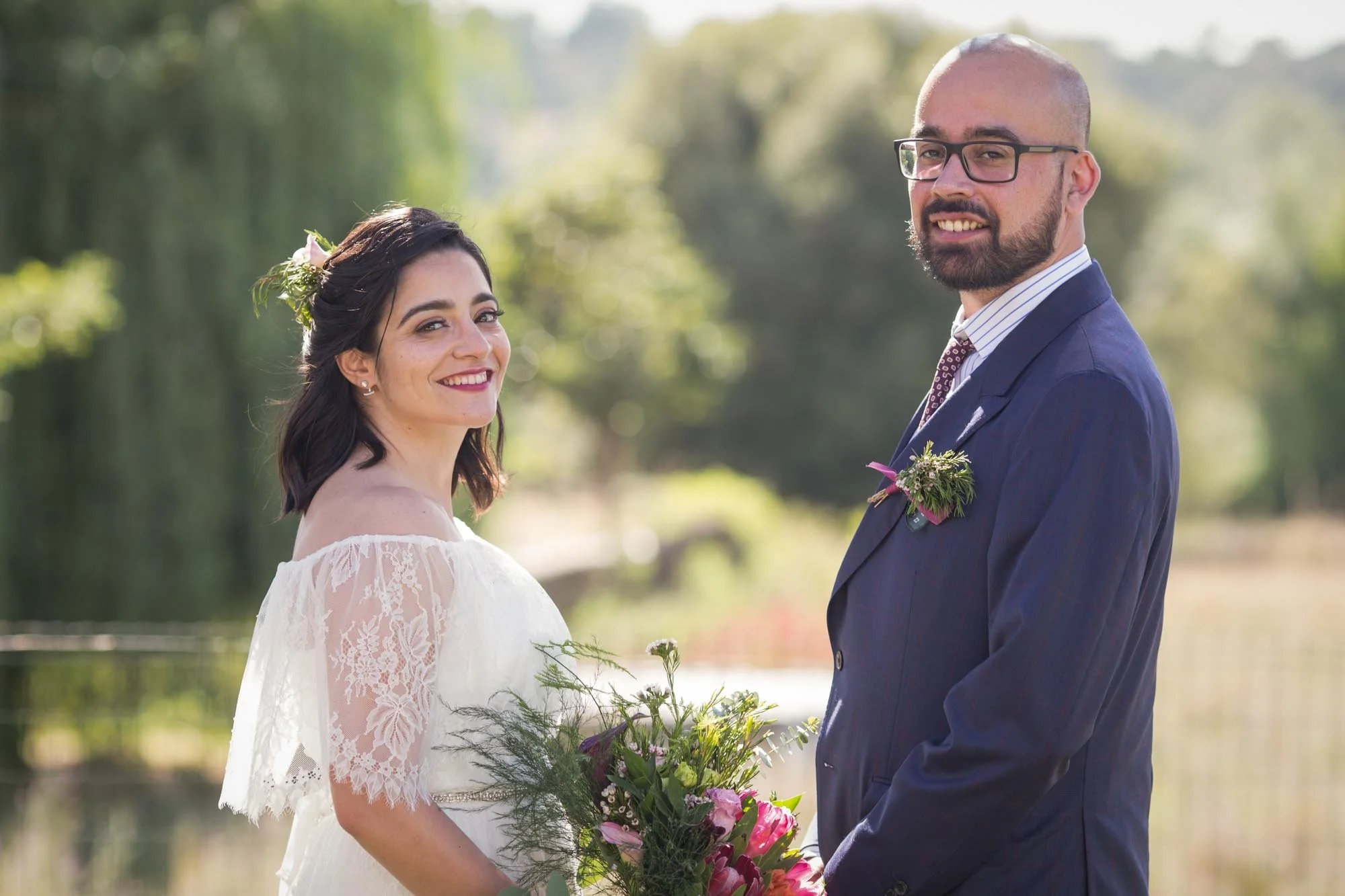 A bride and groom outdoors on a sunny day, smiling and holding a bouquet of flowers, with a blurred green background of trees and water.