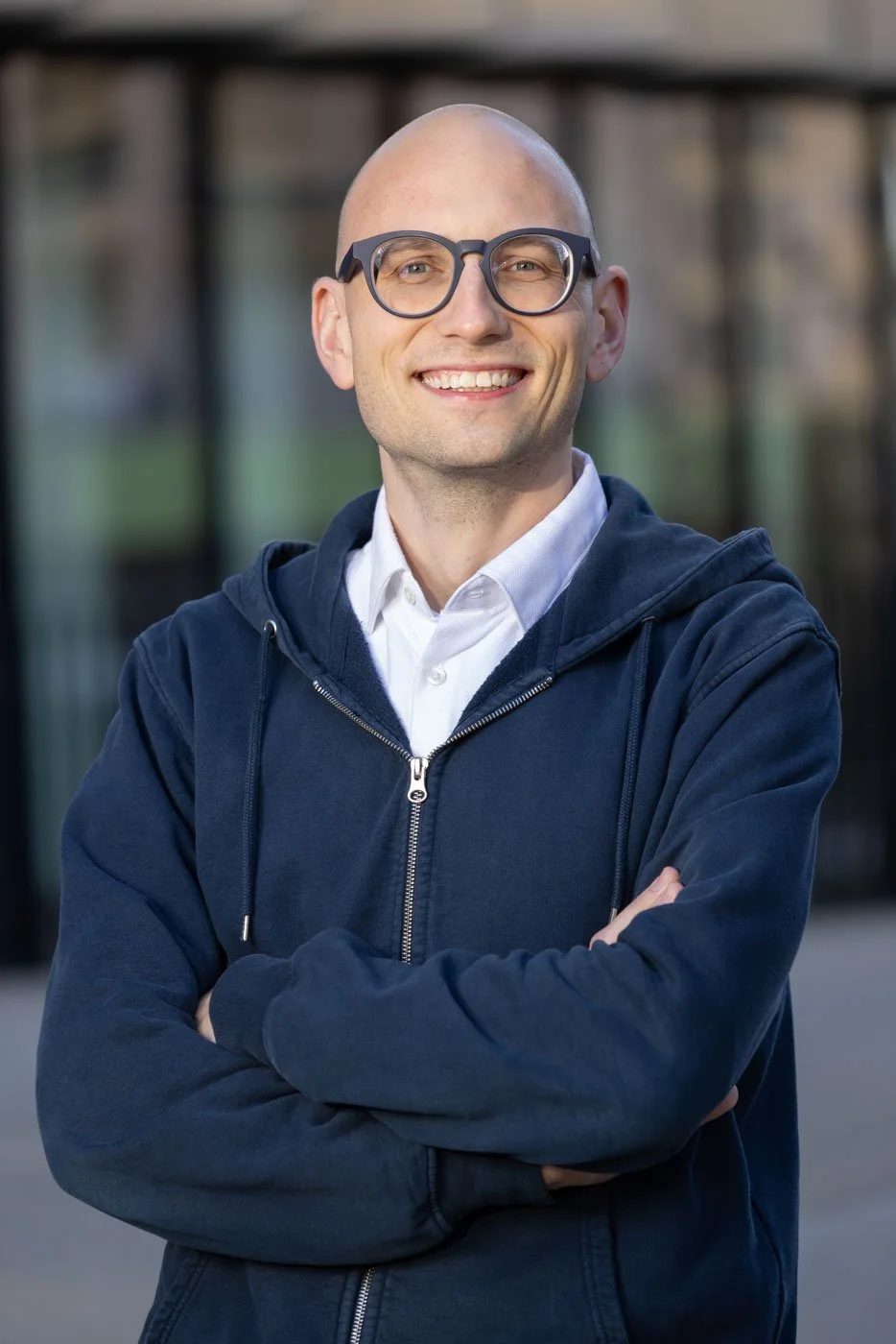 A smiling man with glasses, a bald head, wearing a white shirt and navy blue hoodie, standing outdoors with arms crossed.