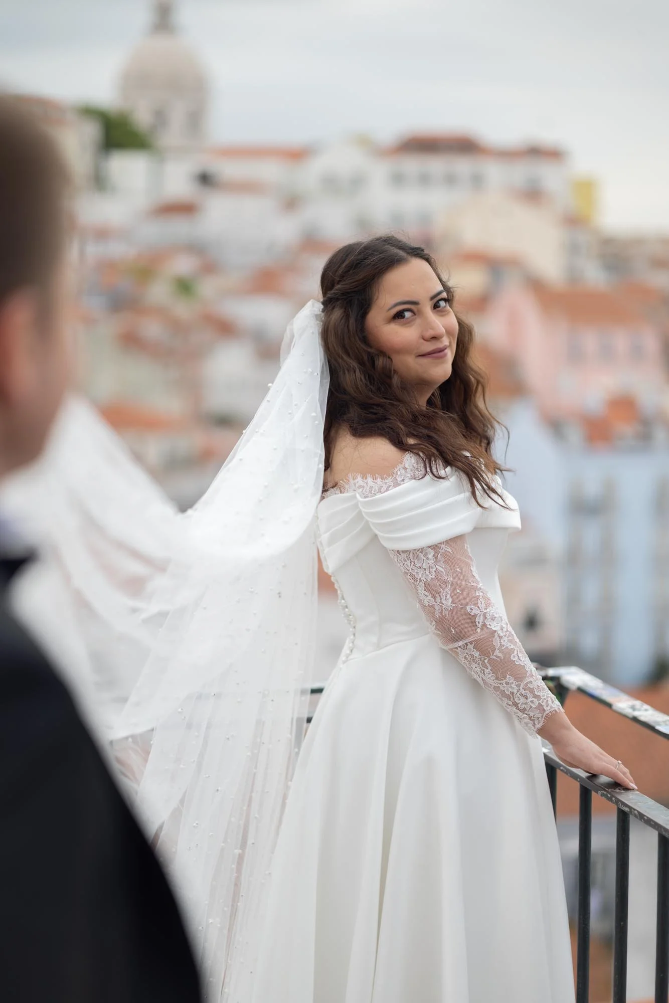 Bridal couple on a balcony, woman's face visible with wedding dress and veil, cityscape background.