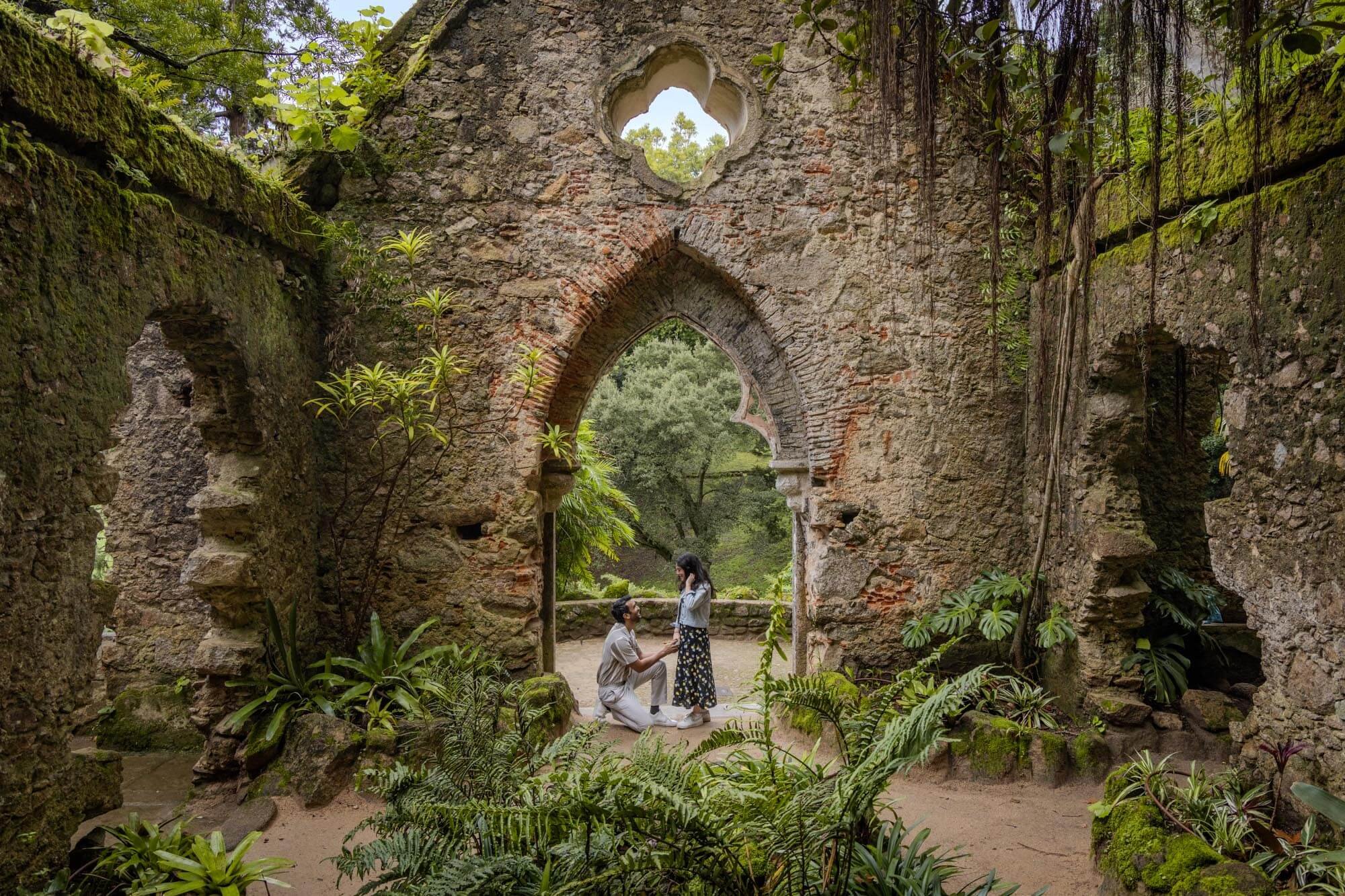 A couple stands in an ancient, ruined stone structure with arched windows, surrounded by lush greenery and plants.