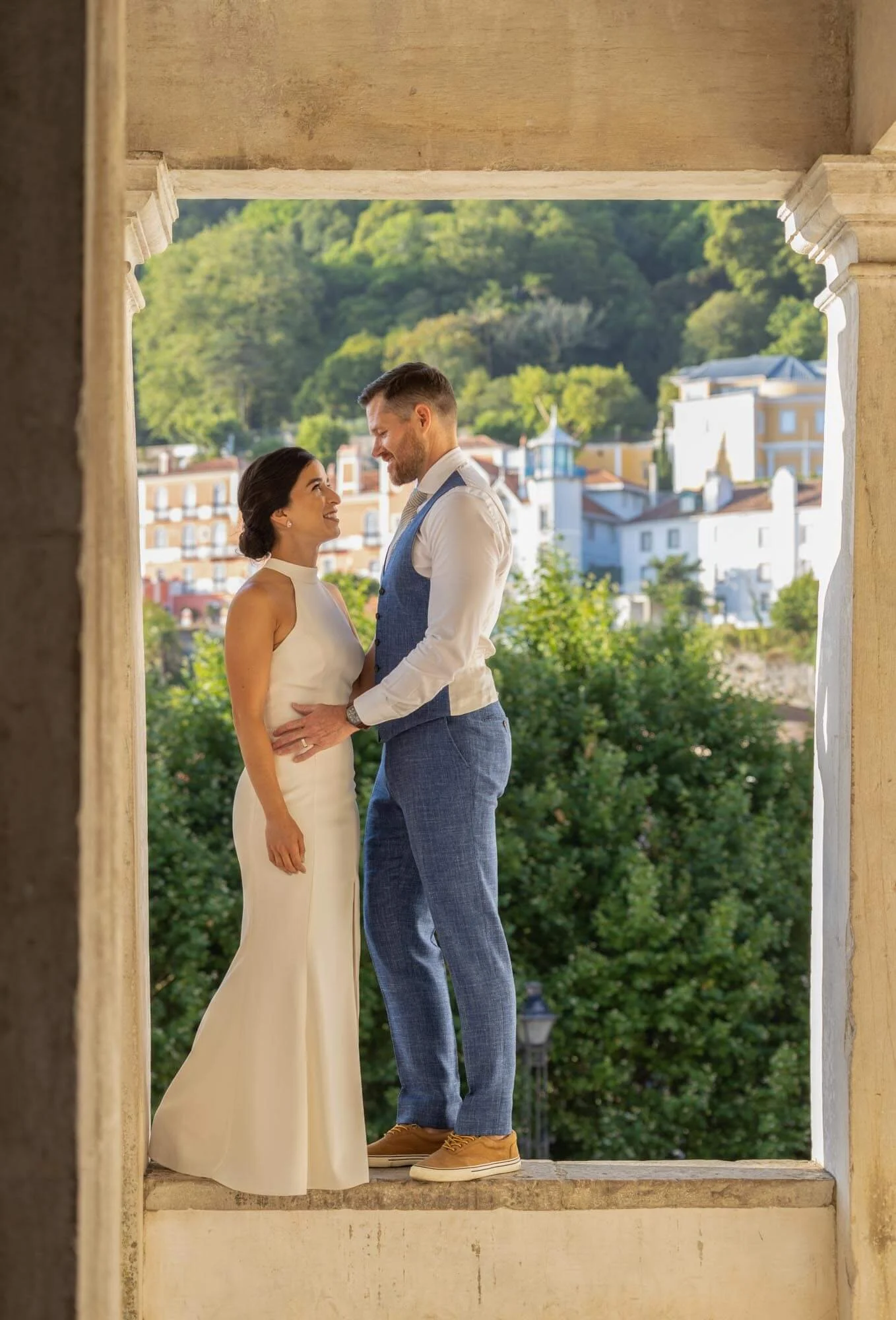 A couple in wedding attire standing on a stone ledge, gazing at each other, with a scenic hillside of Sintra and white buildings in the background.