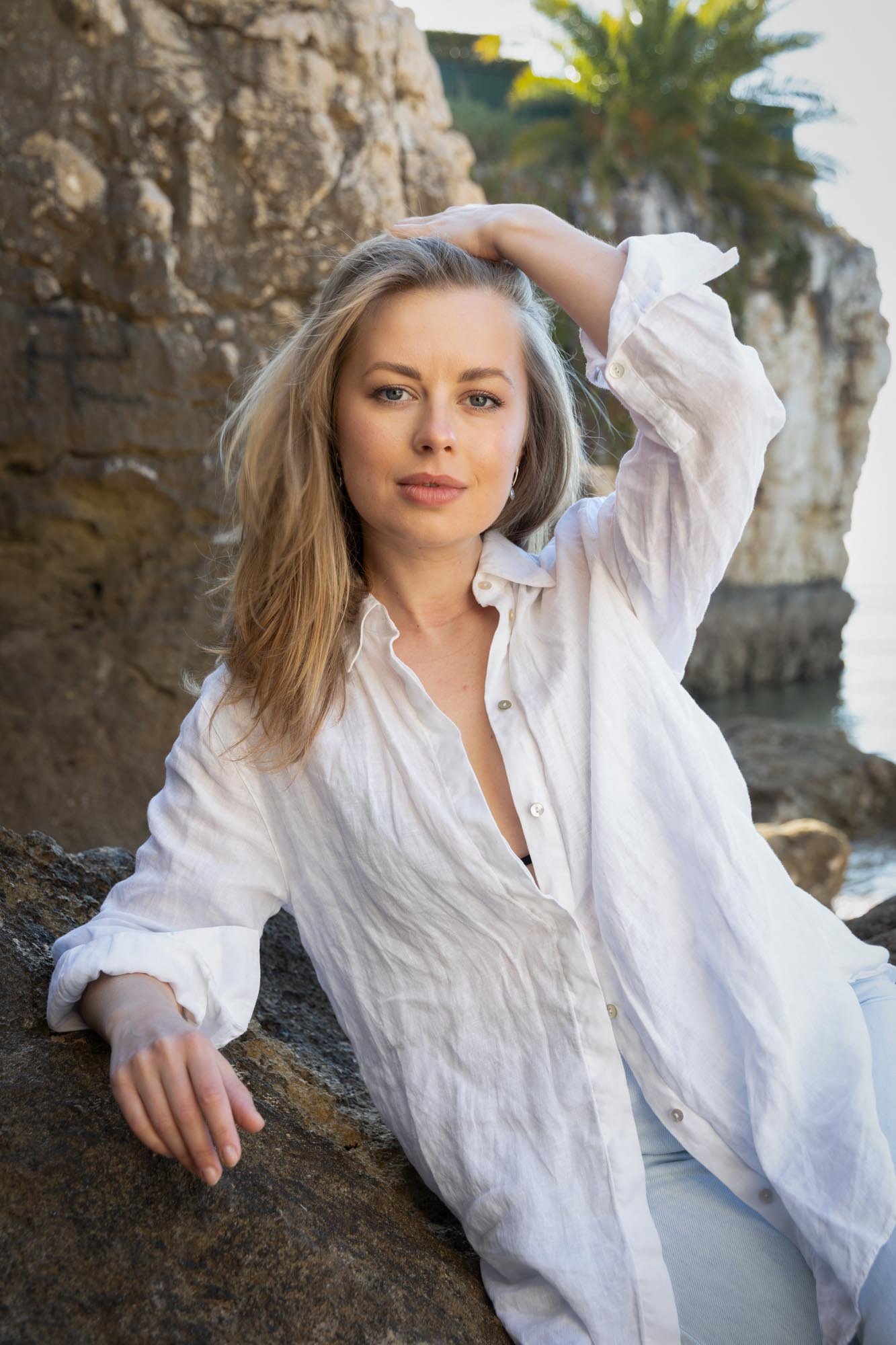 A woman with blonde hair in a white shirt sitting on rocks near water with cliffs in the background, looking at the camera.