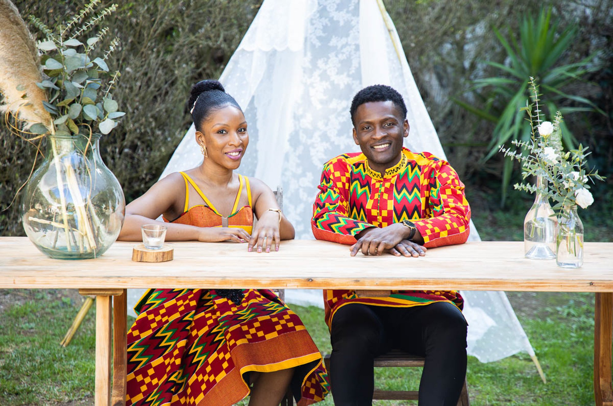 A smiling woman and man sitting at a wooden table outdoors, decorated with glass vases of flowers, greenery, and a white tent in the background.