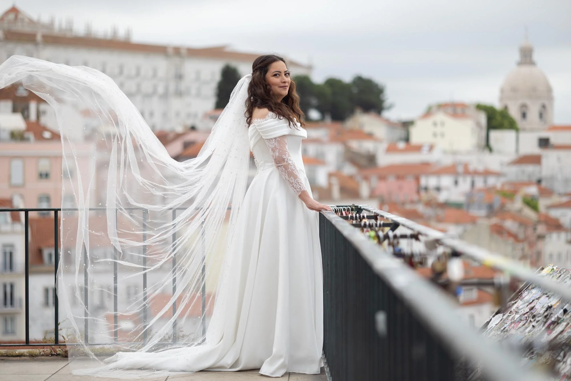A woman in a white wedding dress with lace sleeves standing on a balcony overlooking a city with orange rooftops and a domed building in the background. She has long, wavy brown hair and is smiling slightly while holding onto the railing.