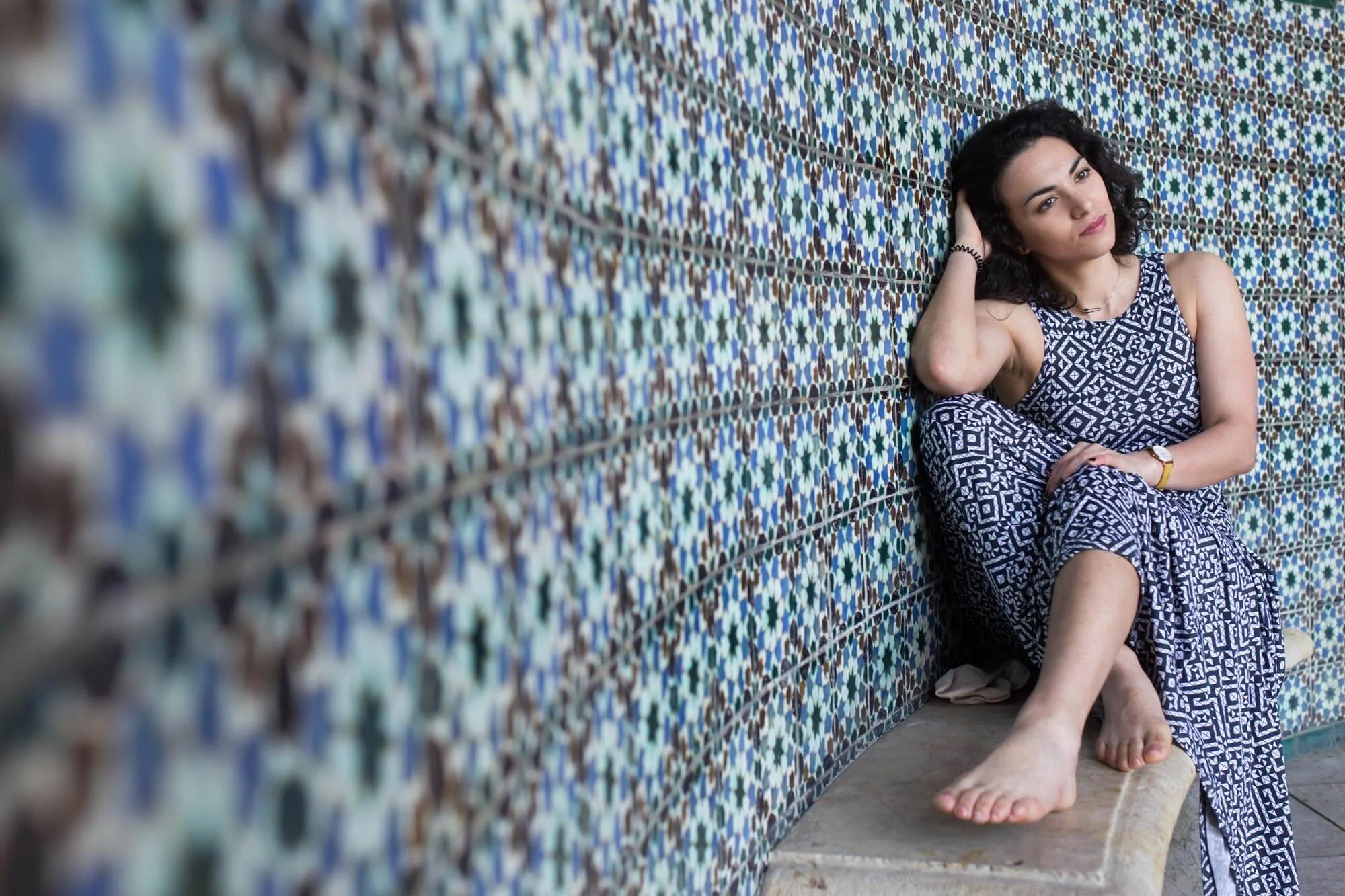 Woman with black curly hair in a patterned dress sitting on a stone bench against a wall covered in colorful Moroccan tile mosaic.
