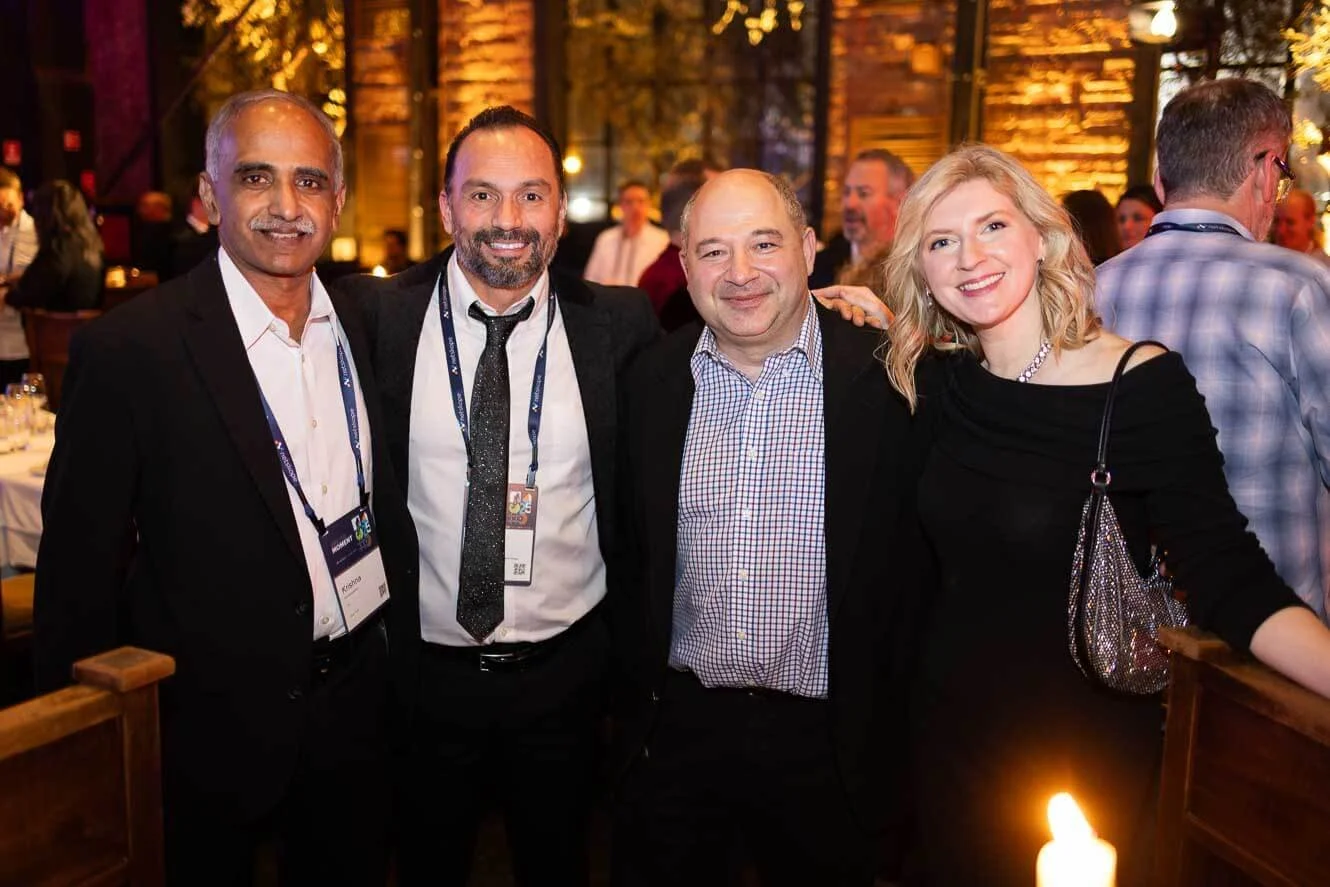 Four people, two men and two women, at a social event, standing together and smiling for the camera in a warmly lit, rustic-style venue with other people in the background.