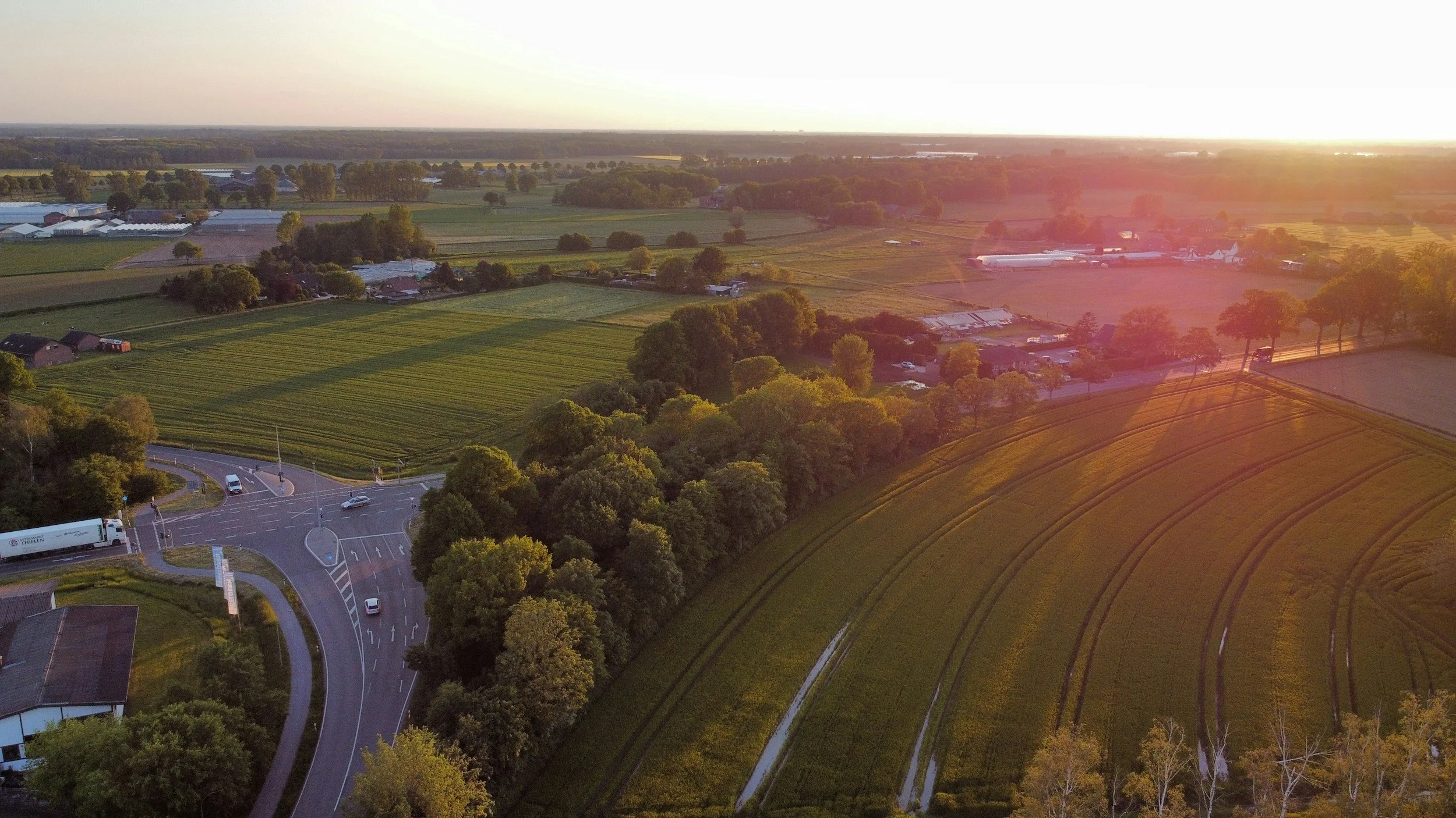 Landschaft mit Straßen, Feldern, Bäumen und Häusern bei Sonnenuntergang