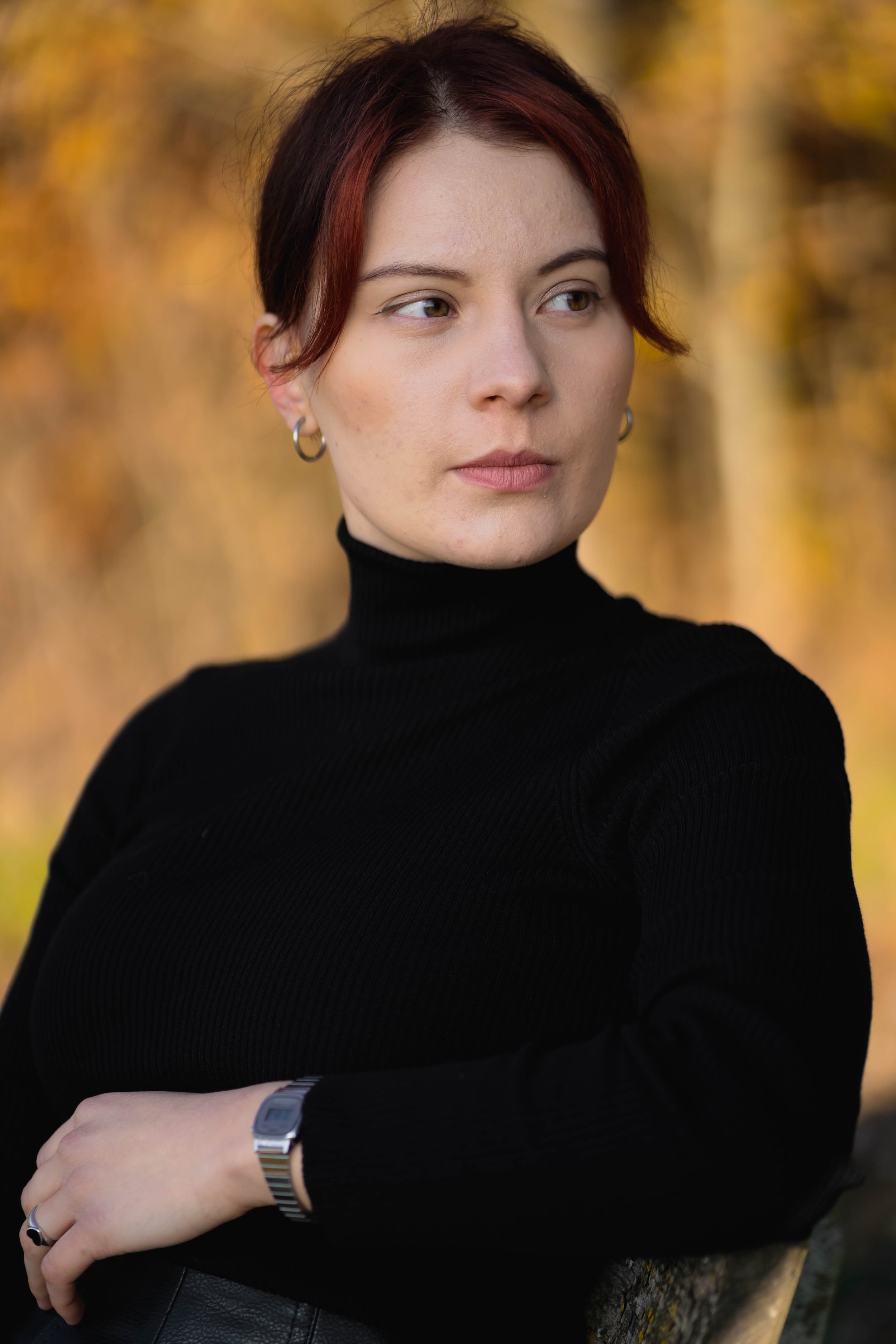 Junge Frau mit braunen Haaren und Ohrringen trägt schwarzen Rollkragenpullover, blickt nach rechts, im Hintergrund Herbstblätter in warmen Farbtönen.