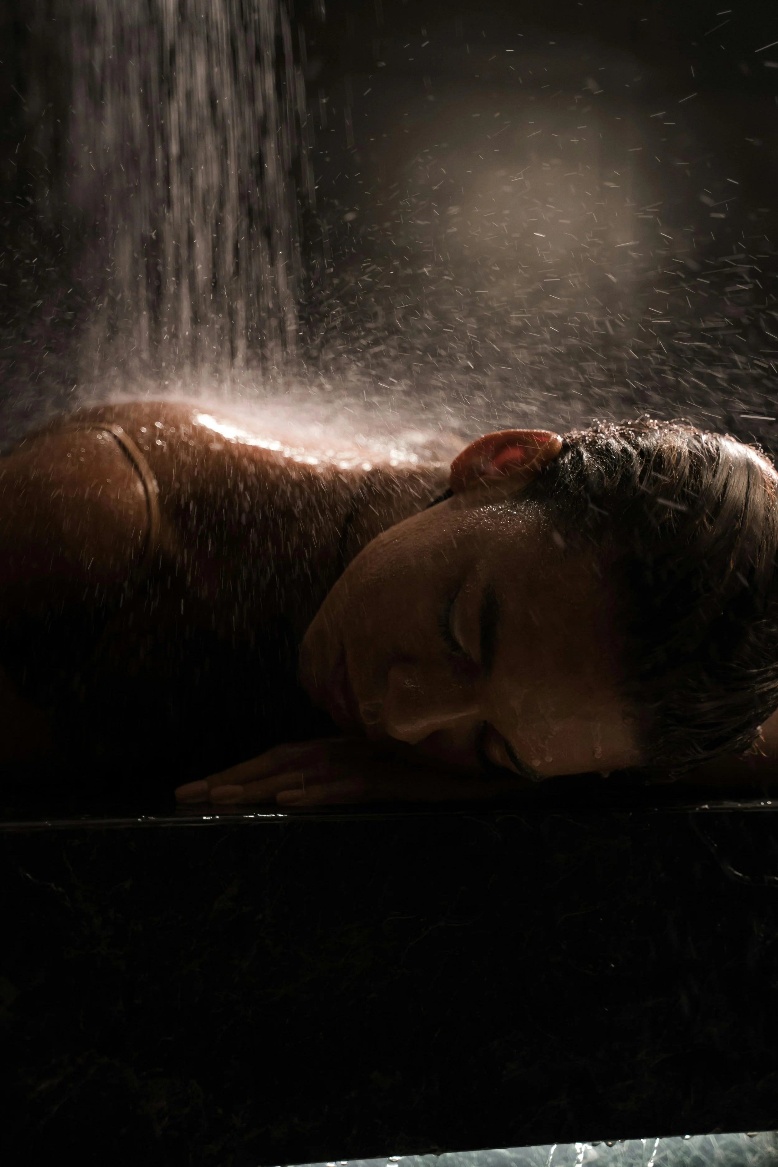 Une femme repose dans une baignoire ou un espace de bain, sous une pluie artificielle ou une douche, dans une ambiance sombre et relaxante.