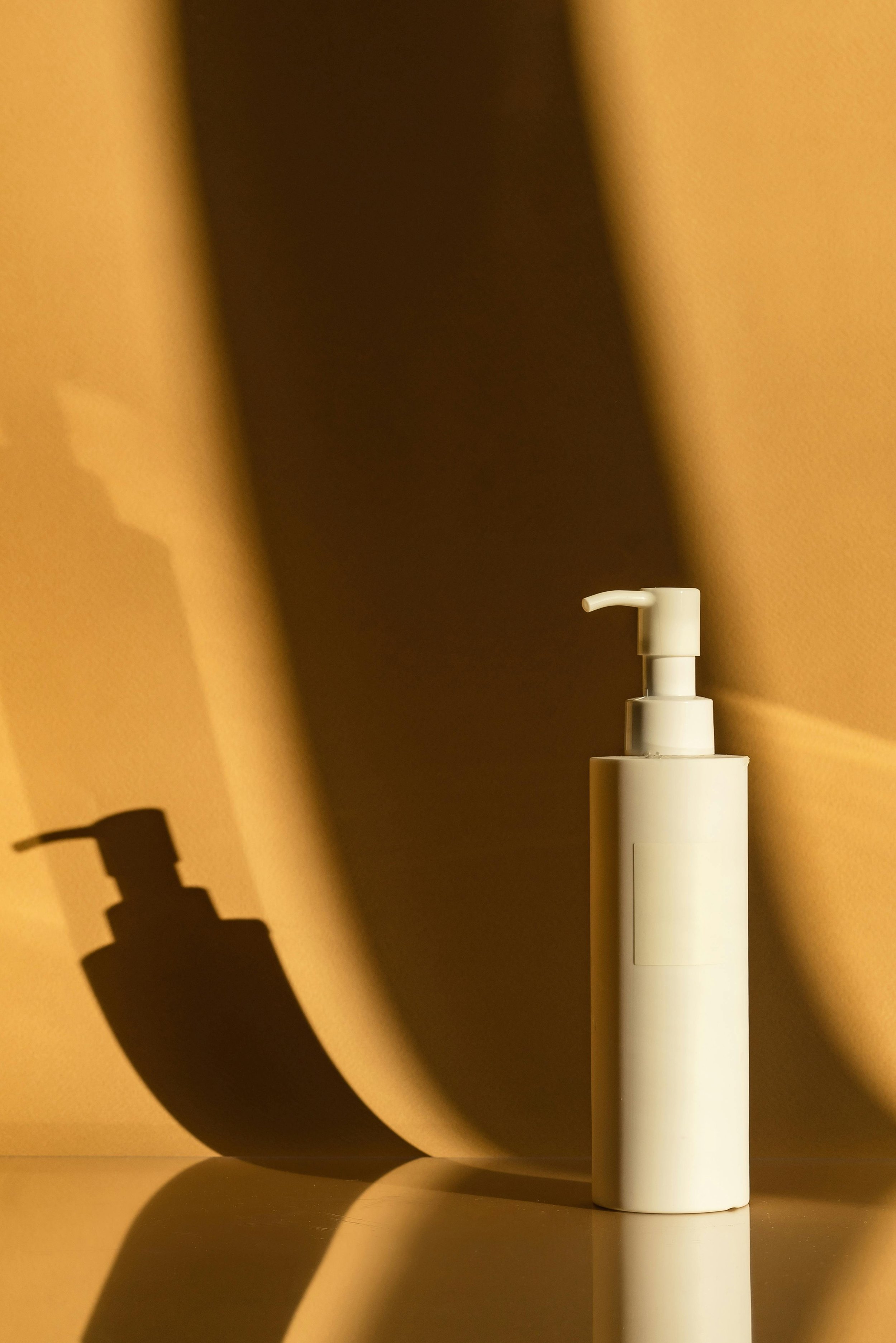 Une bouteille blanche de savon avec pompe, posée sur une surface claire, crée une ombre noire décorative sur un mur beige en arrière-plan.