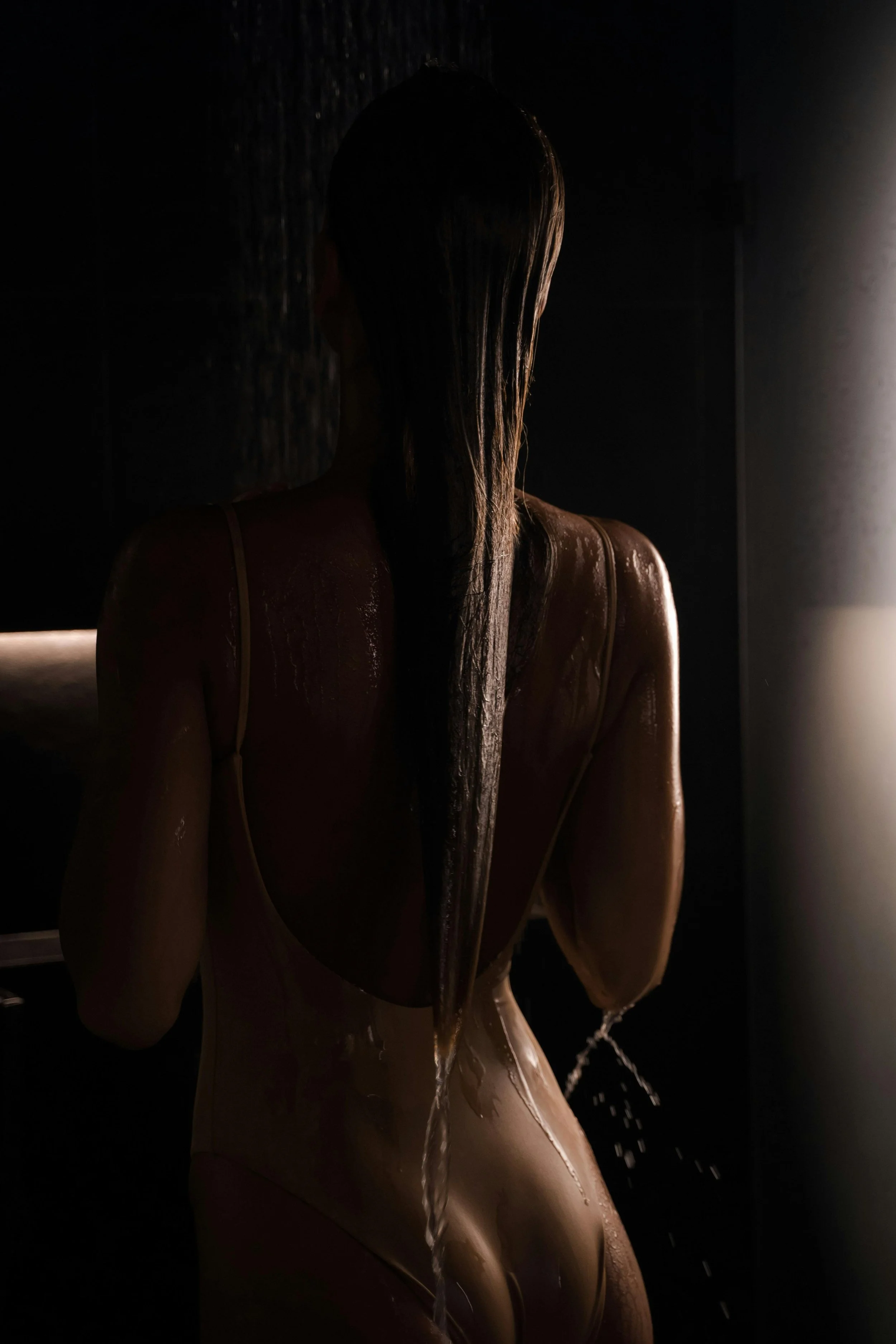 Une personne vue de dos sous une lumière tamisée, les cheveux mouillés, portant une tenue moulante, dans une pièce sombre.
