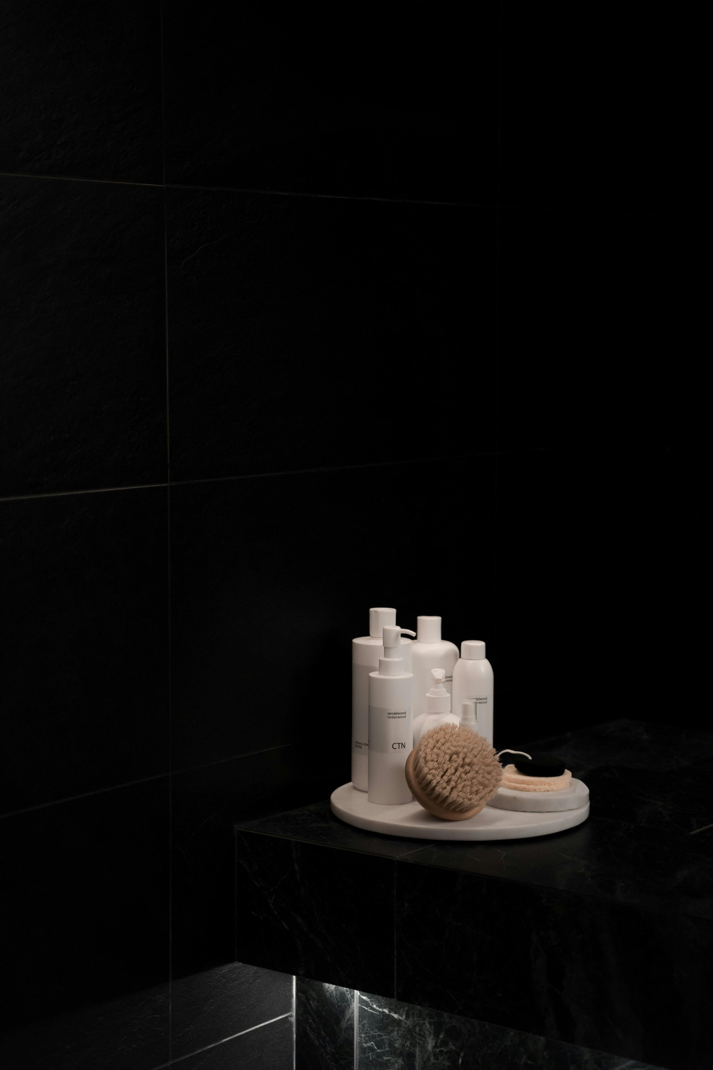 Produits de soins personnels blancs et une brosse sur un plateau blanc dans une salle de bain sombre.