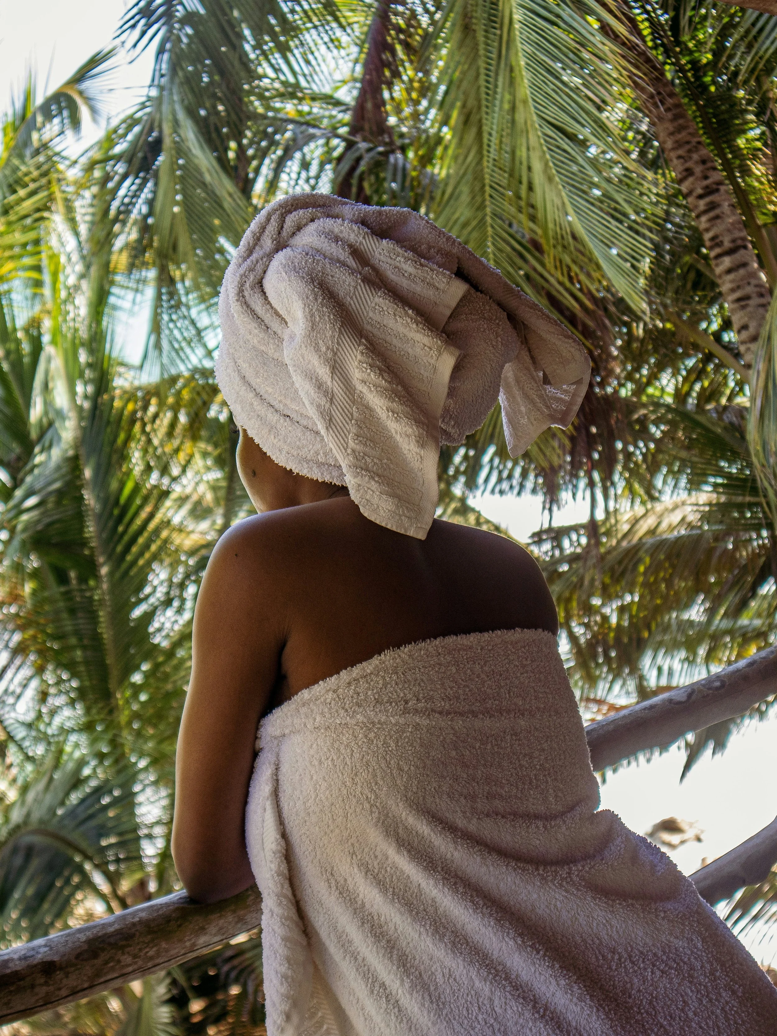 Une personne vue de dos, enveloppée dans une serviette blanche, portant une serviette enroulée autour de la tête, se tenant près d'une barrière en bois dans un environnement verdoyant tropical avec des palmier.