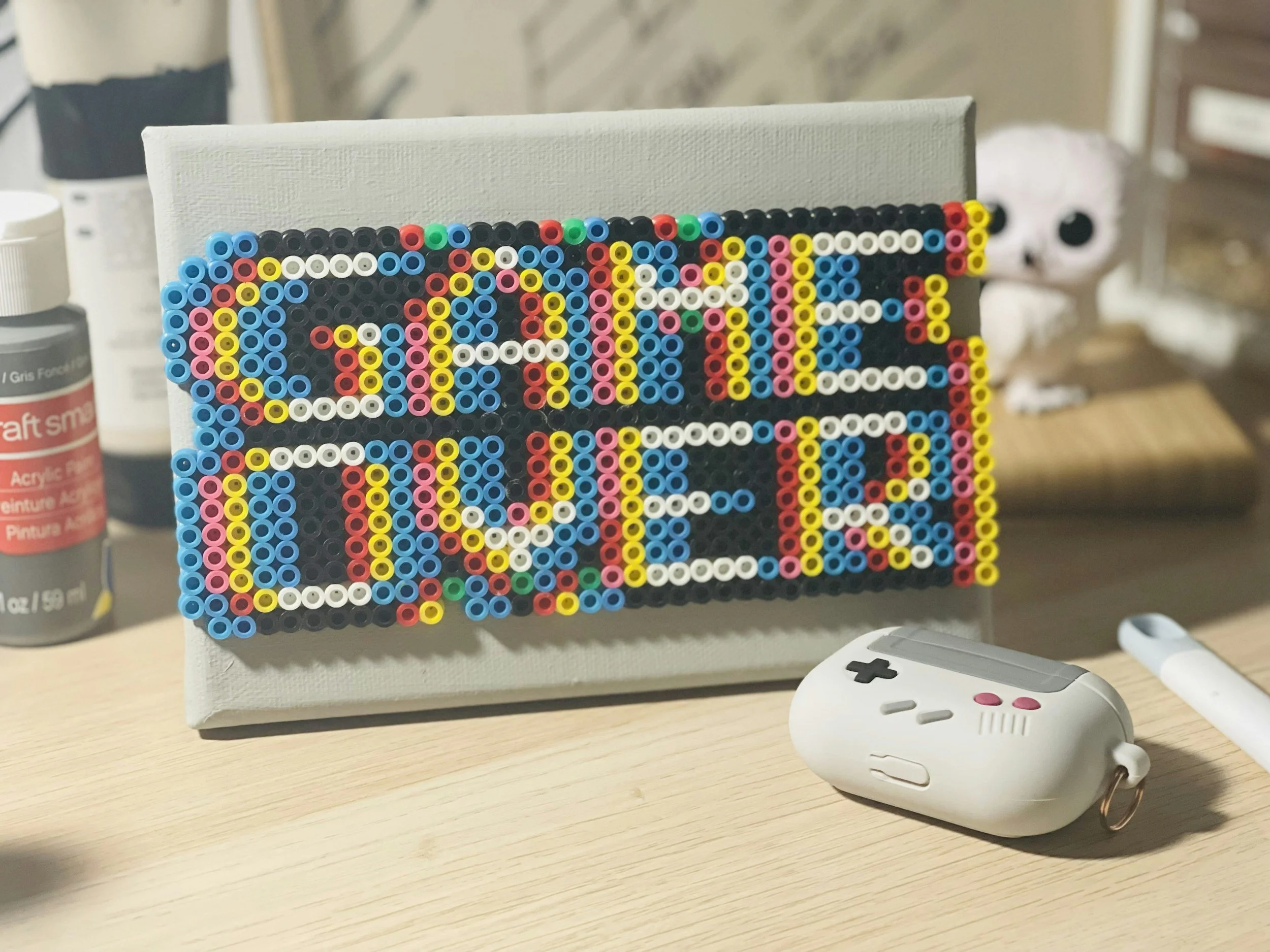 A pixel art piece made from colored beads spelling out the words "GAME OVER" on a white canvas, with a gaming controller wireless mouse and bottles in the background on a wooden surface. Don't let anyone say game over on your relationship.