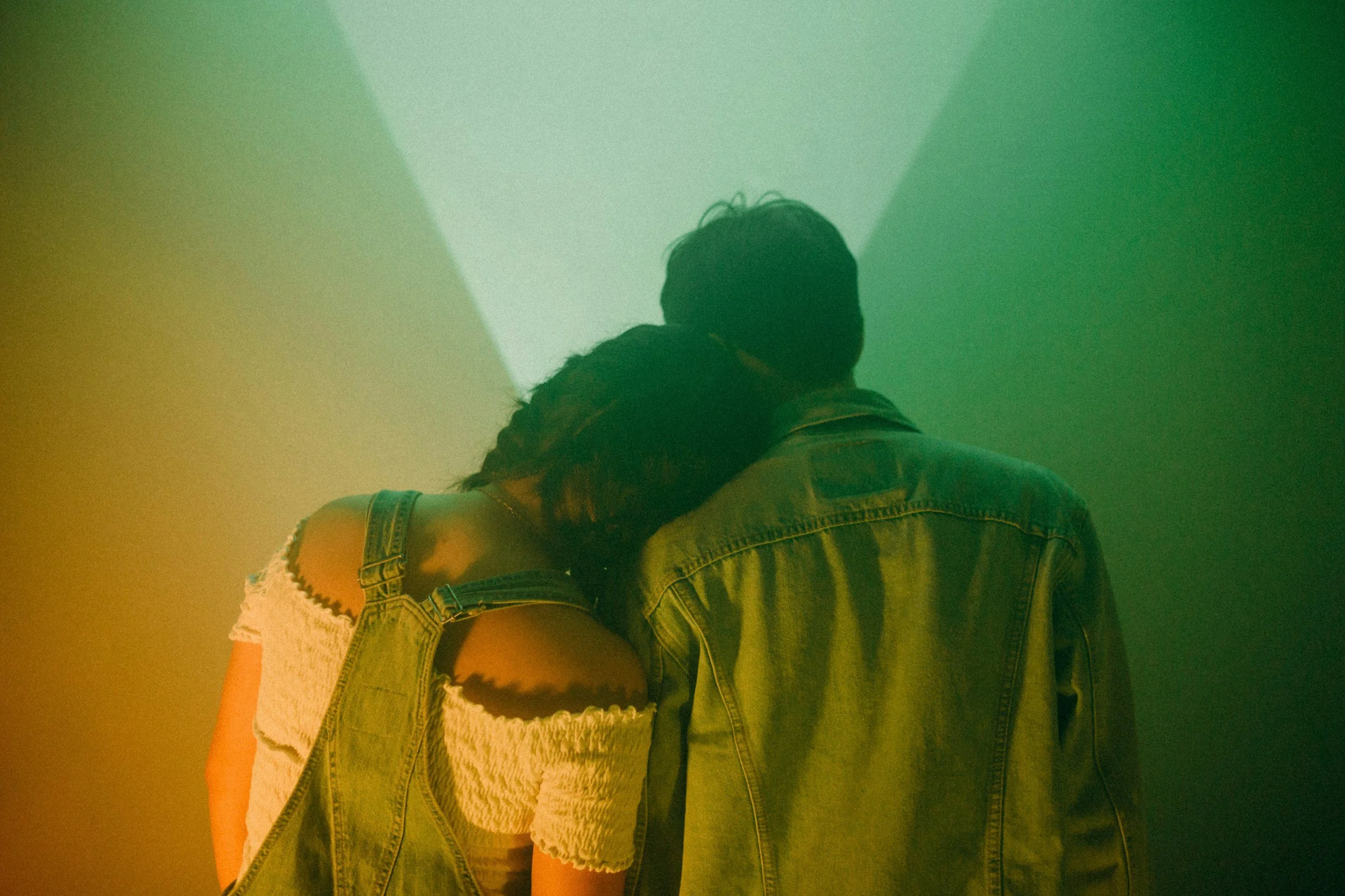 A couple stands with their backs to the camera, the woman resting her head on the man's shoulder against a colorful background with green and yellow hues.