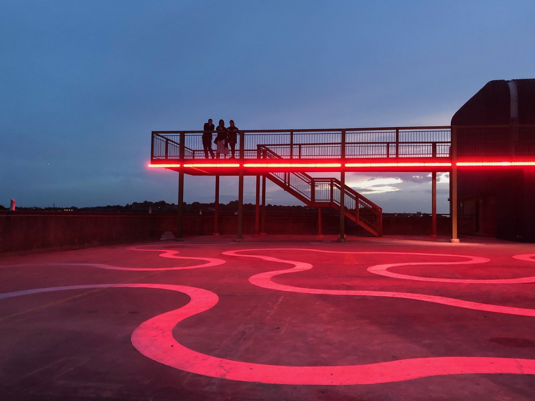 A rooftop deck with three people standing on a balcony, illuminated by red LED strip lights, against a dusk sky with clouds and cityscape in the distance.