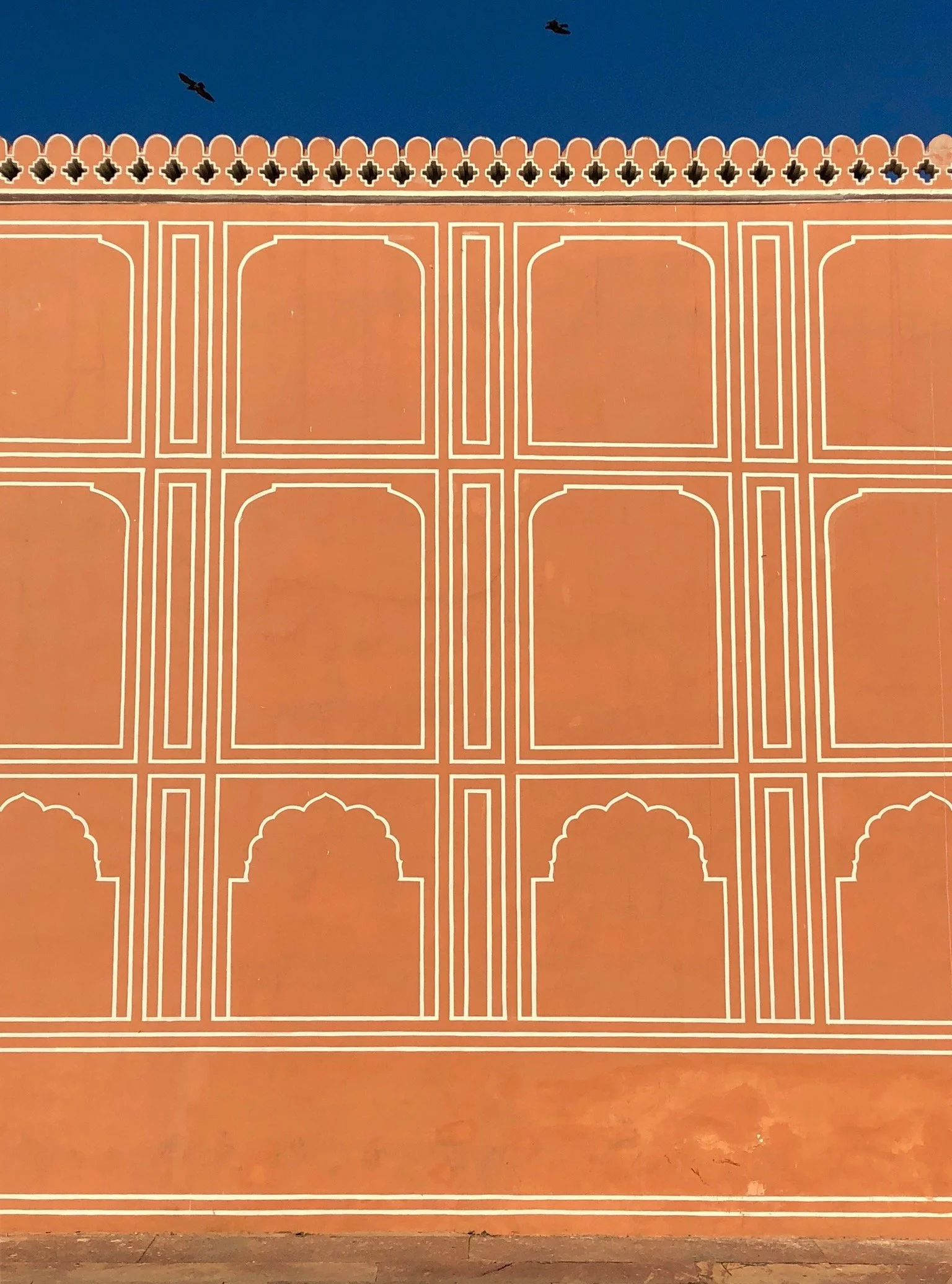 A wall painted in orange with white decorative patterns, featuring a series of arches and rectangular designs. The sky above is clear blue with two birds flying.