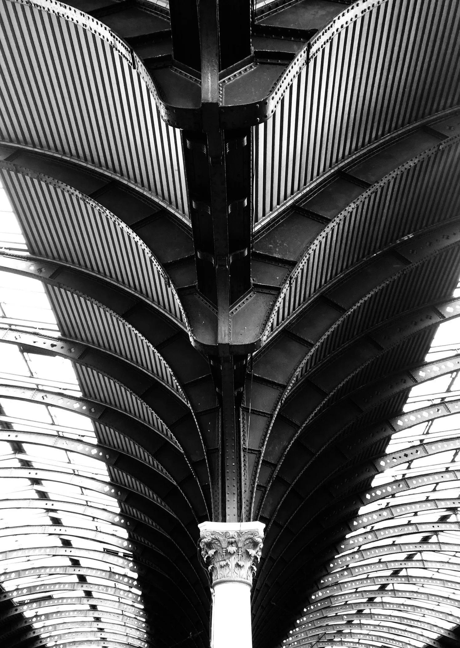 Black and white photo of an ornate architectural ceiling with curved metal beams and a column with Greek-style capitals.
