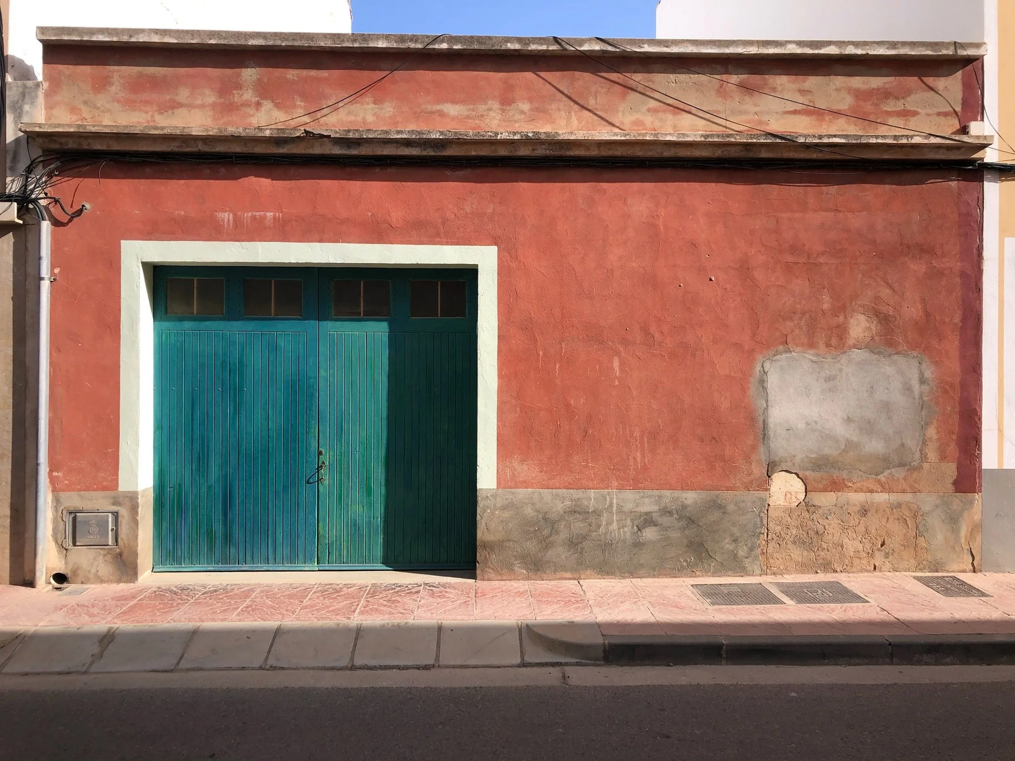 A weathered red building with a closed teal garage door and small windows above it. The building has patches of peeling paint and cracks along the wall, with a concrete sidewalk and street in front.