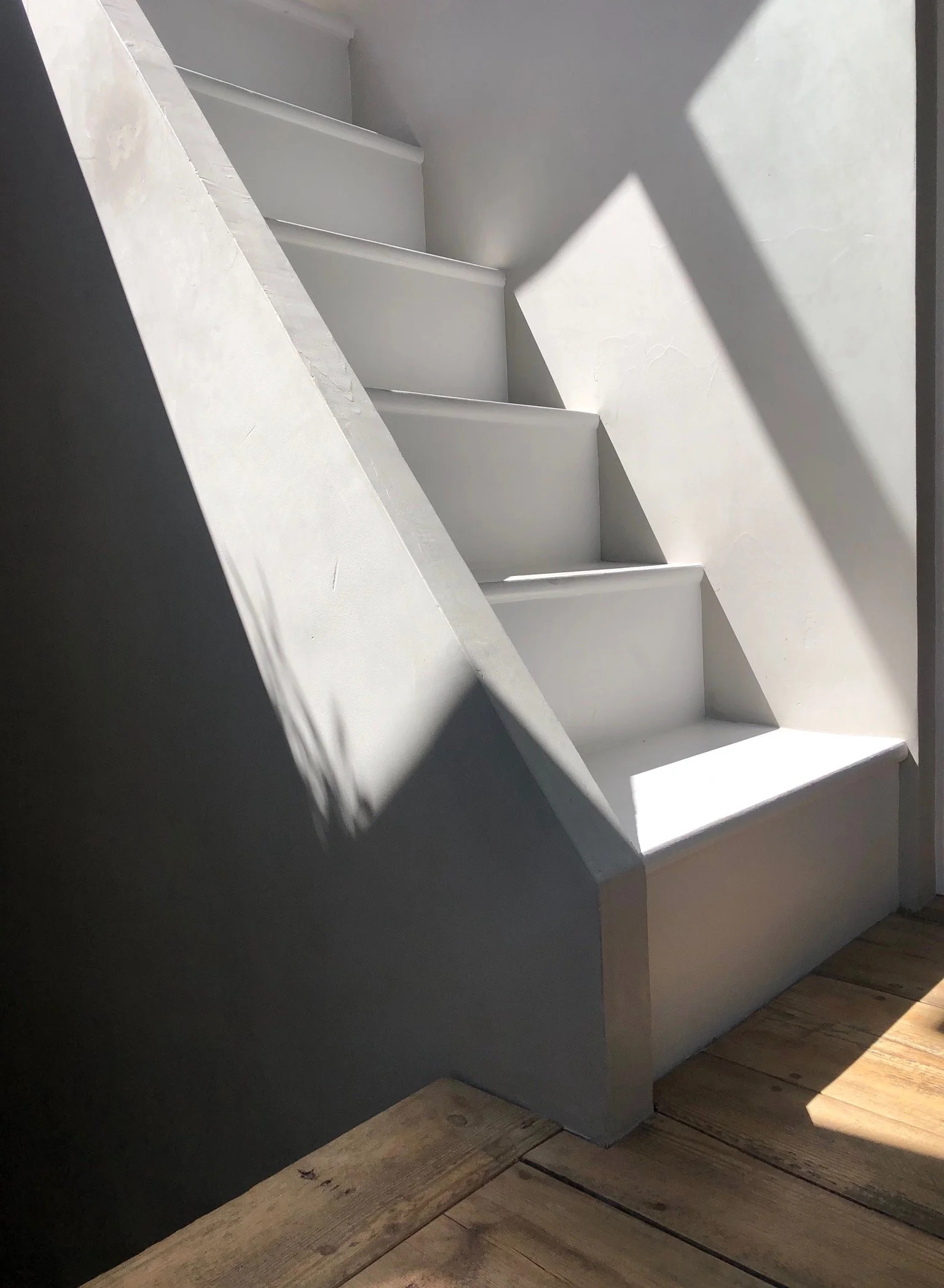 White staircase with shadows cast on the wall and wooden floorboards.