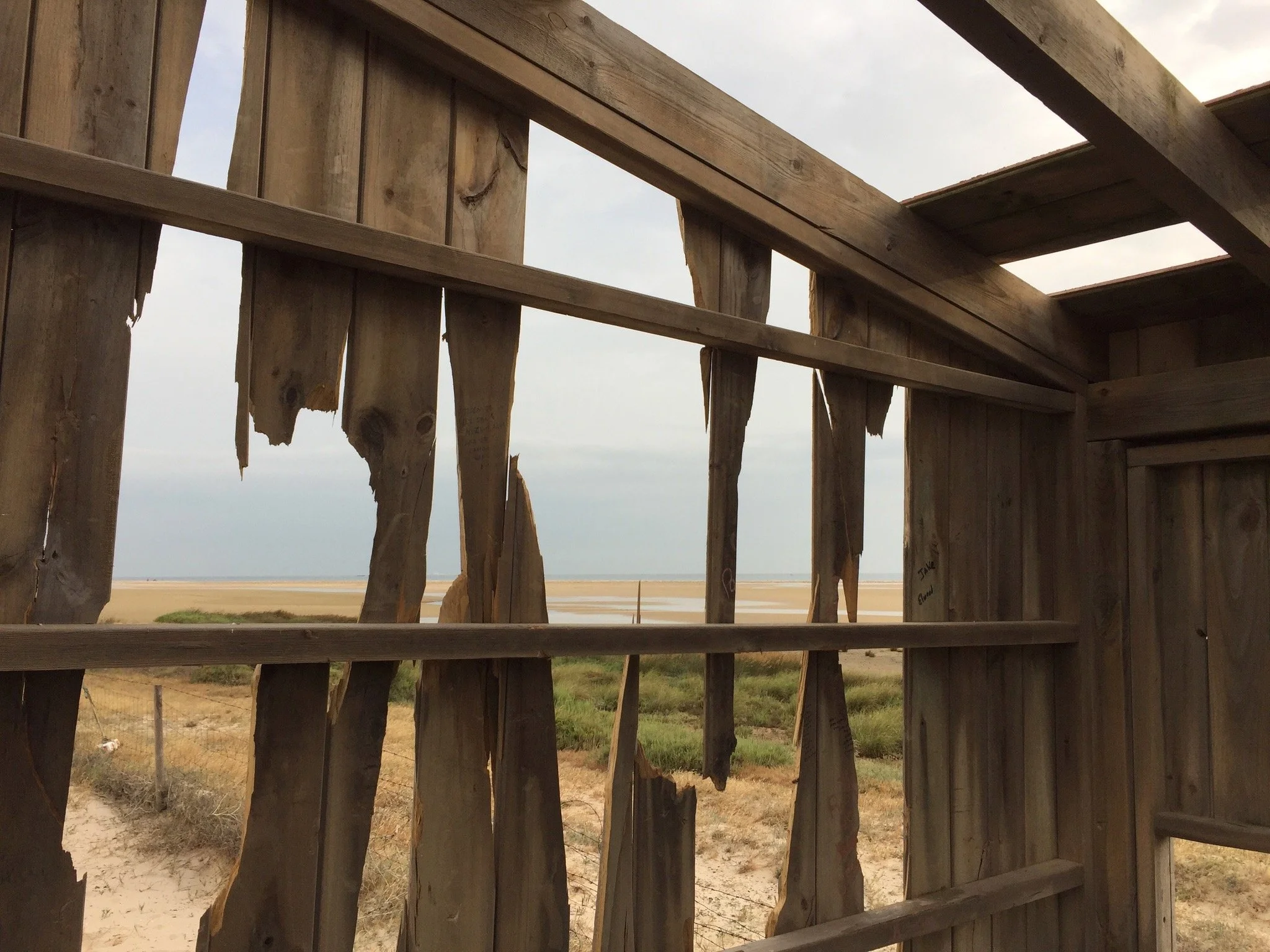 View through a broken wooden fence overlooking a sandy beach and water, with a cloudy sky.