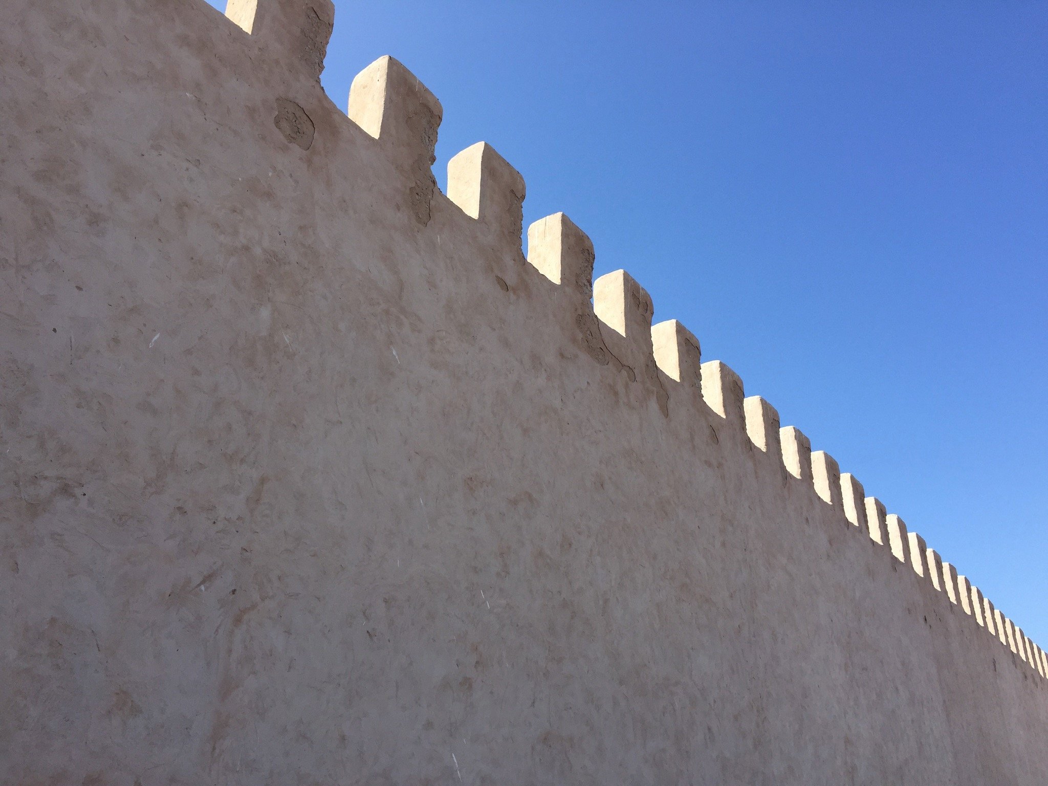 Close-up of a beige adobe wall with crenellations at the top, against a clear blue sky.