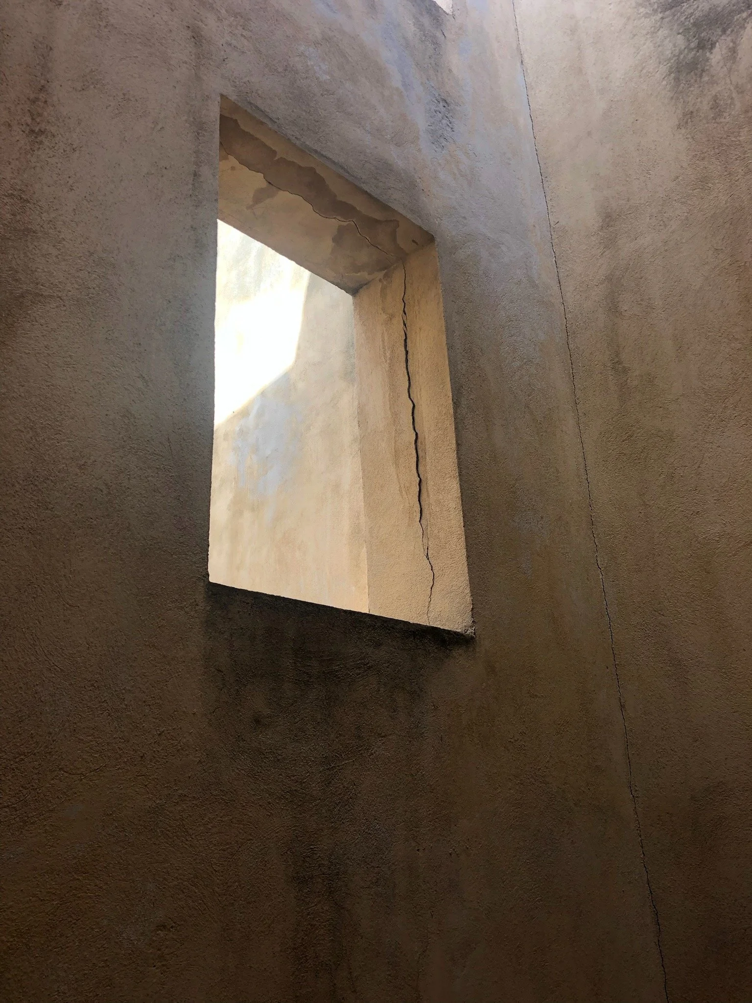 An interior wall with a rectangular opening, showing a crack running vertically along the wall, with sunlight coming through the opening.