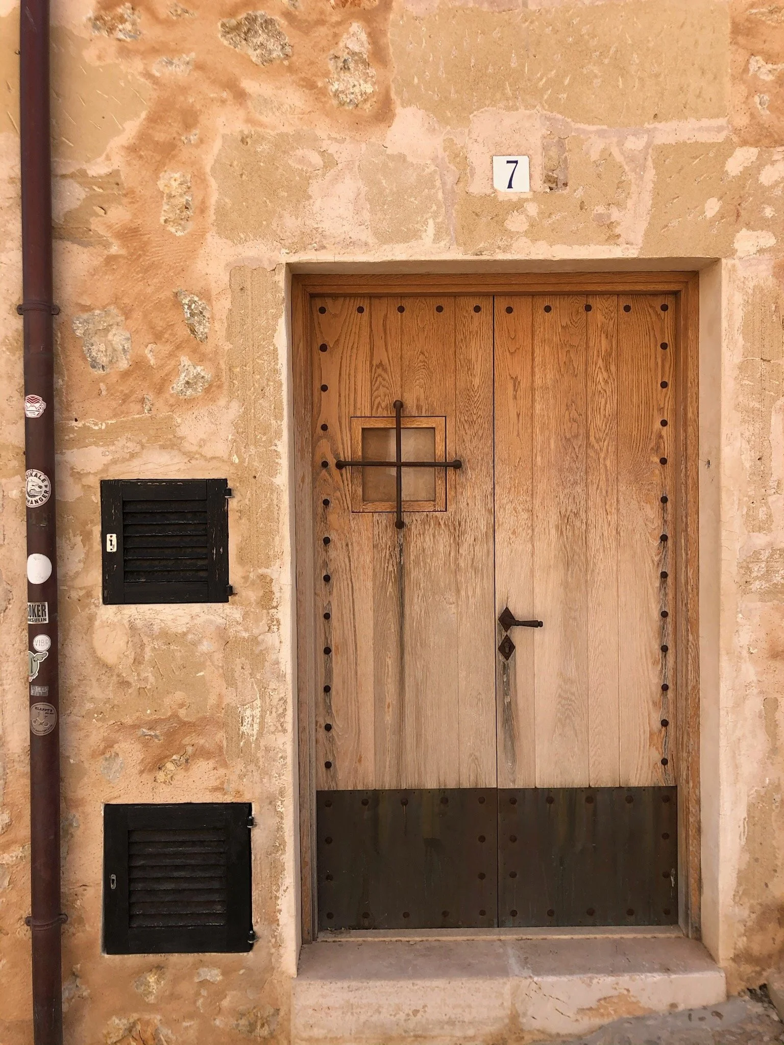 Wooden door with black metal accents, a small square window, and a handle, set within a weathered stone wall with the number 7 above it.
