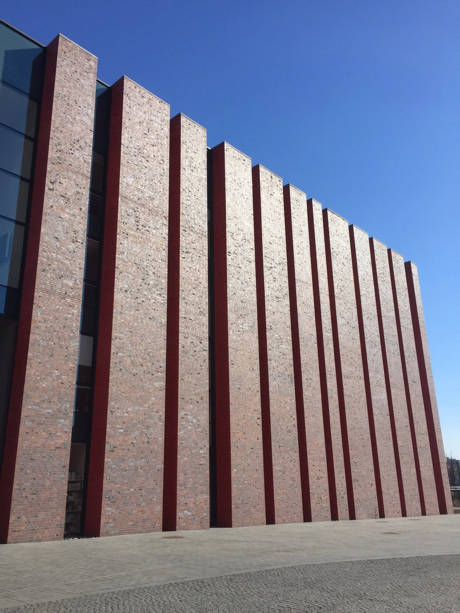 Exterior view of a modern multi-story brick building with vertical red accents, large glass windows on the left, and a clear blue sky above.