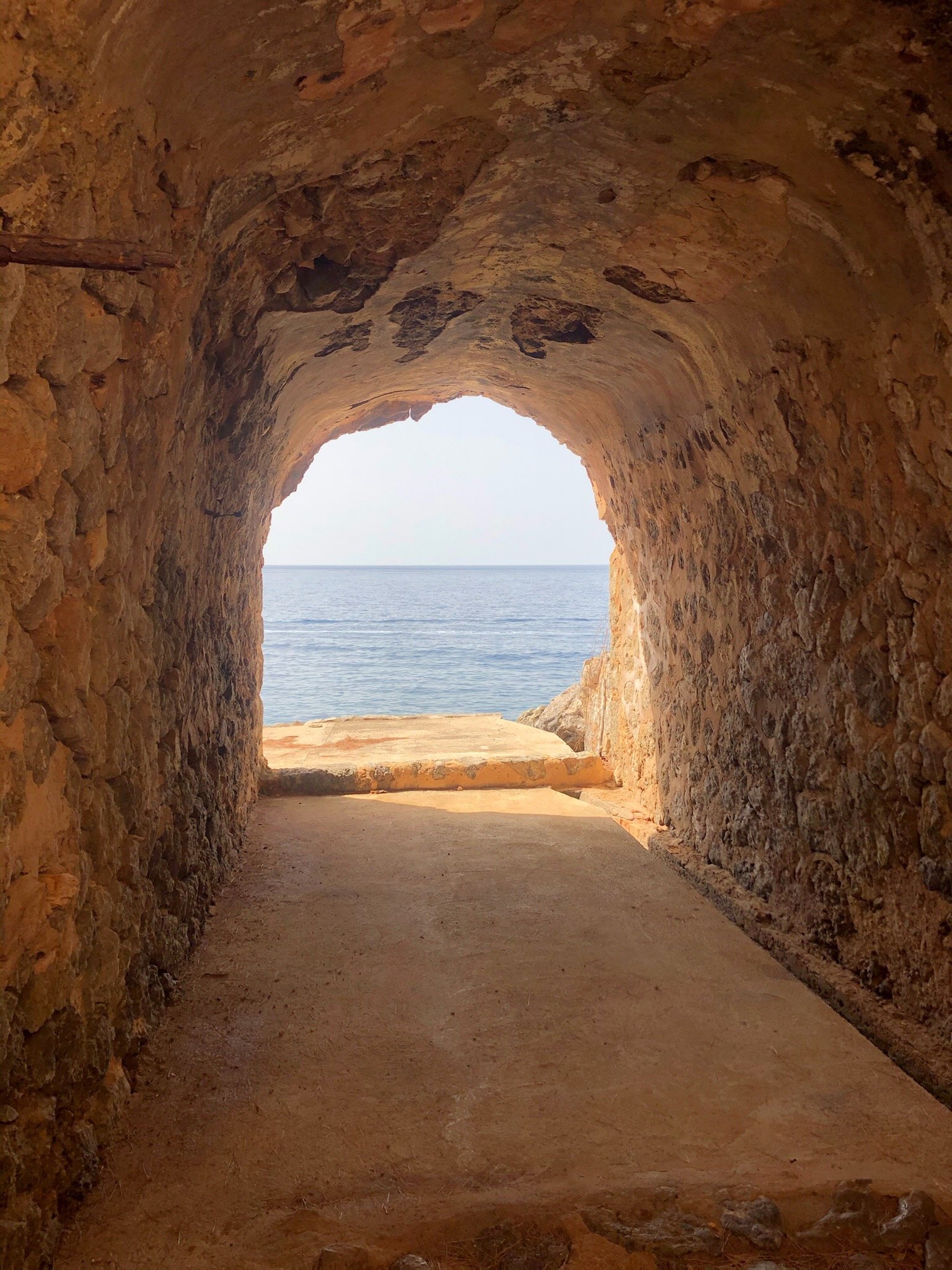 View from inside a stone tunnel opening to the ocean with a clear sky and calm water in the distance.