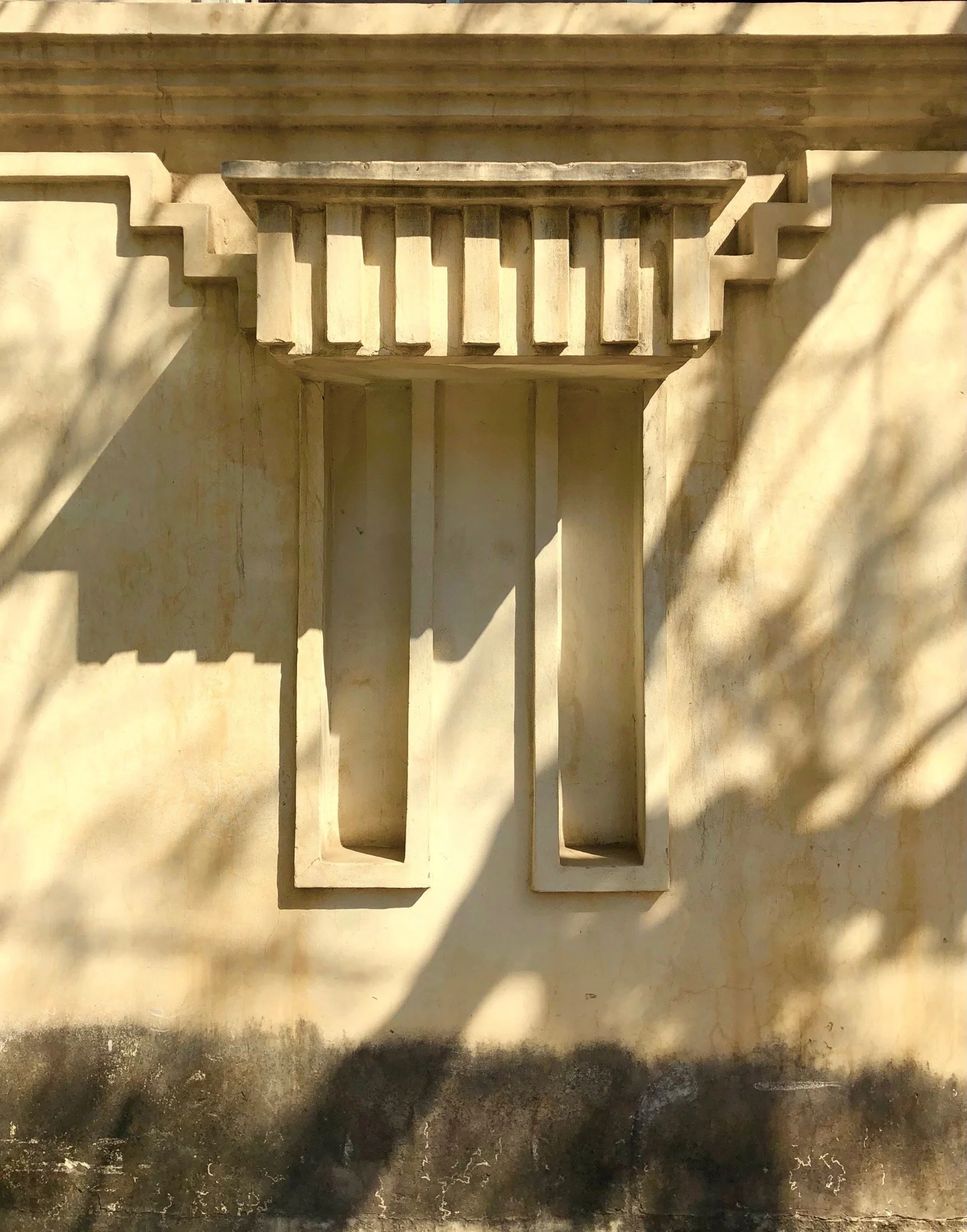 Decorative architectural feature on a wall with shadows and sunlight, resembling a stylized face or mask.
