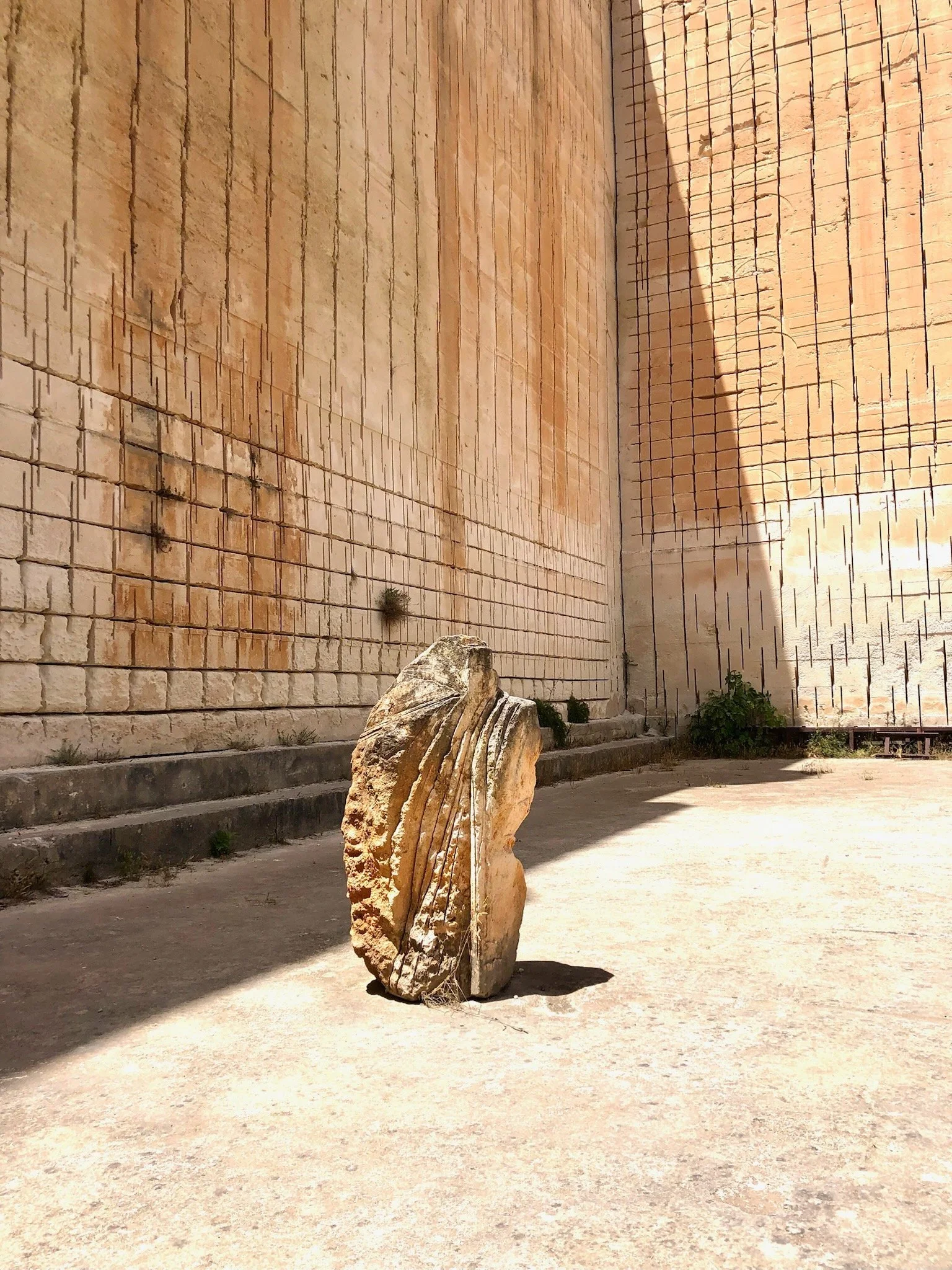 A large rock with carved lines stands on a sunlit concrete ground in front of tall beige stone walls with small plants growing at the base.