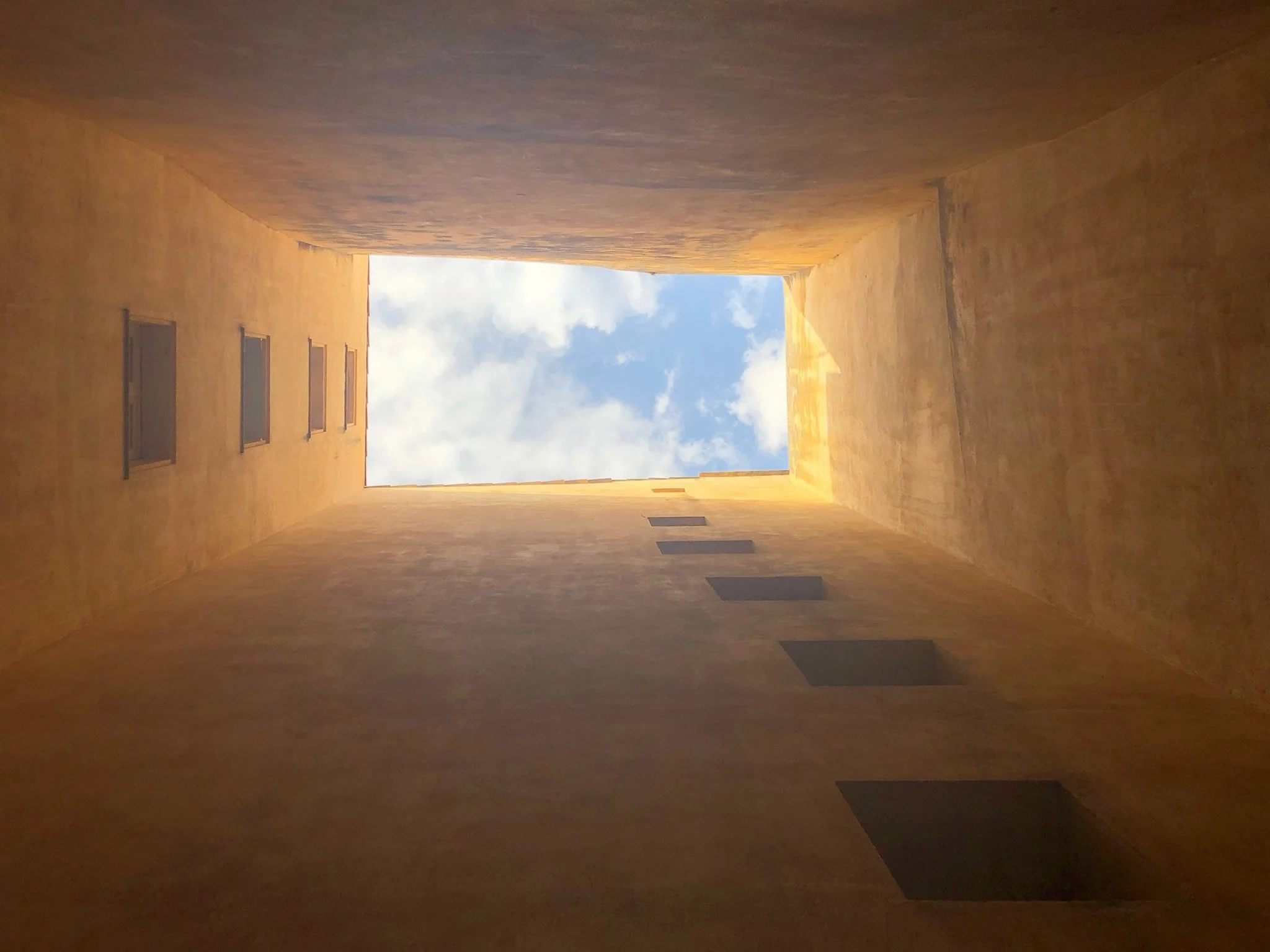 Looking up from the bottom of a rectangular courtyard towards the sky through an opening above. The courtyard walls are yellowish with rectangular windows on one side and small square openings on the other.