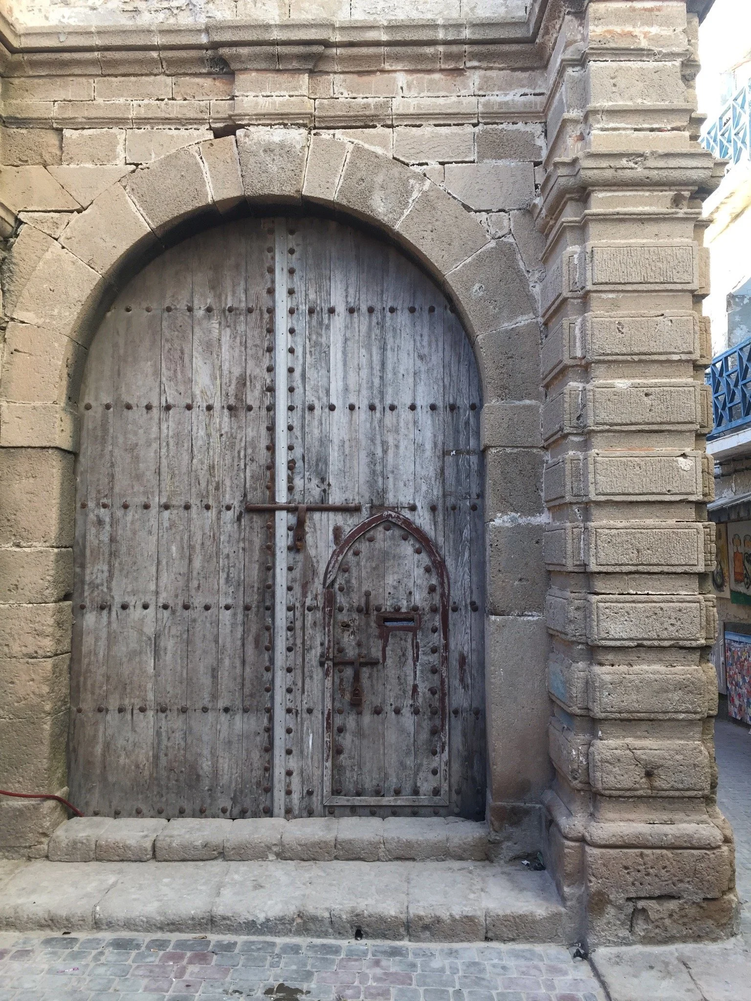 Old wooden double door with iron studs and a small mail slot, set in a stone archway on a city street.