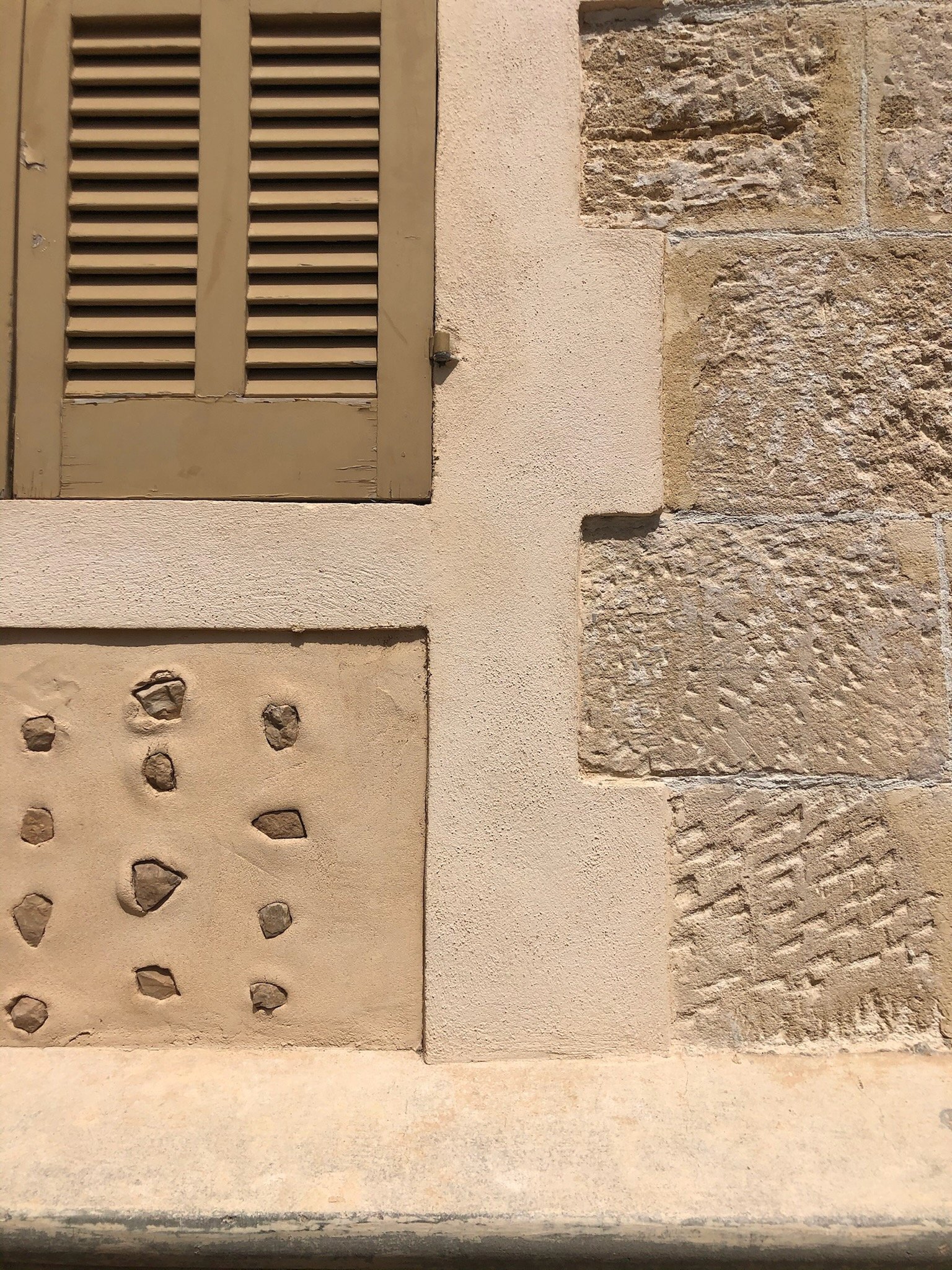 Close-up of a building exterior featuring a beige wooden window shutter, a textured beige wall next to a rough stone section, and a concrete ledge at the bottom.