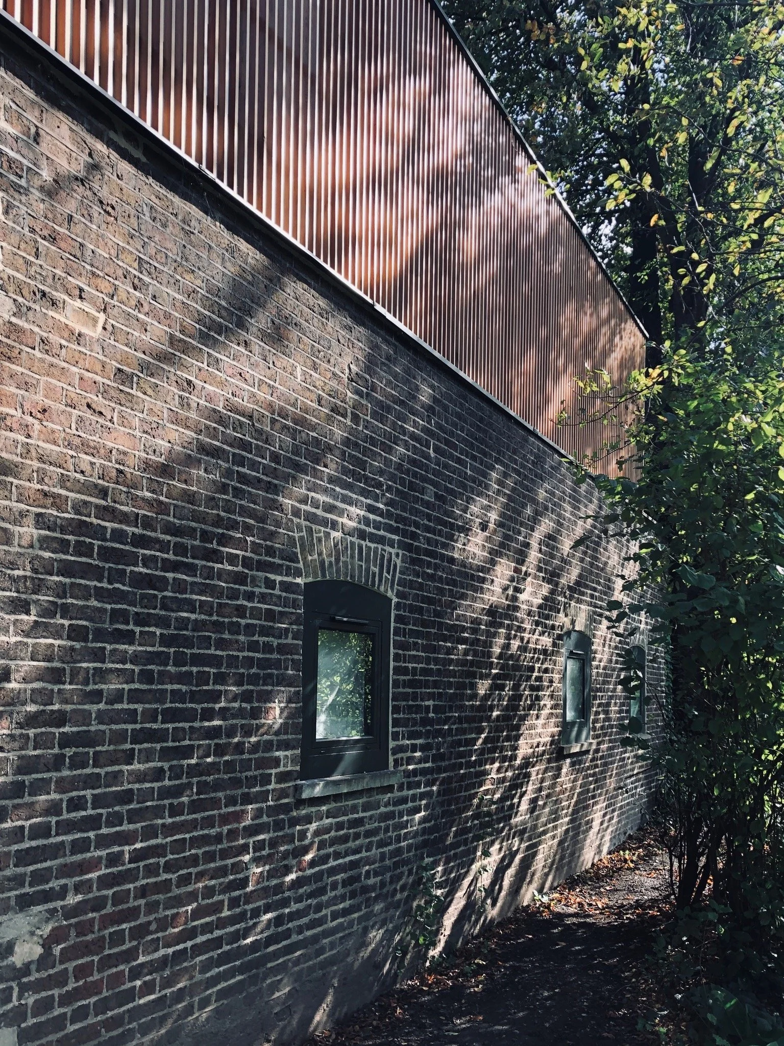 Side of a brick building with two small windows and tree shadows on the wall.