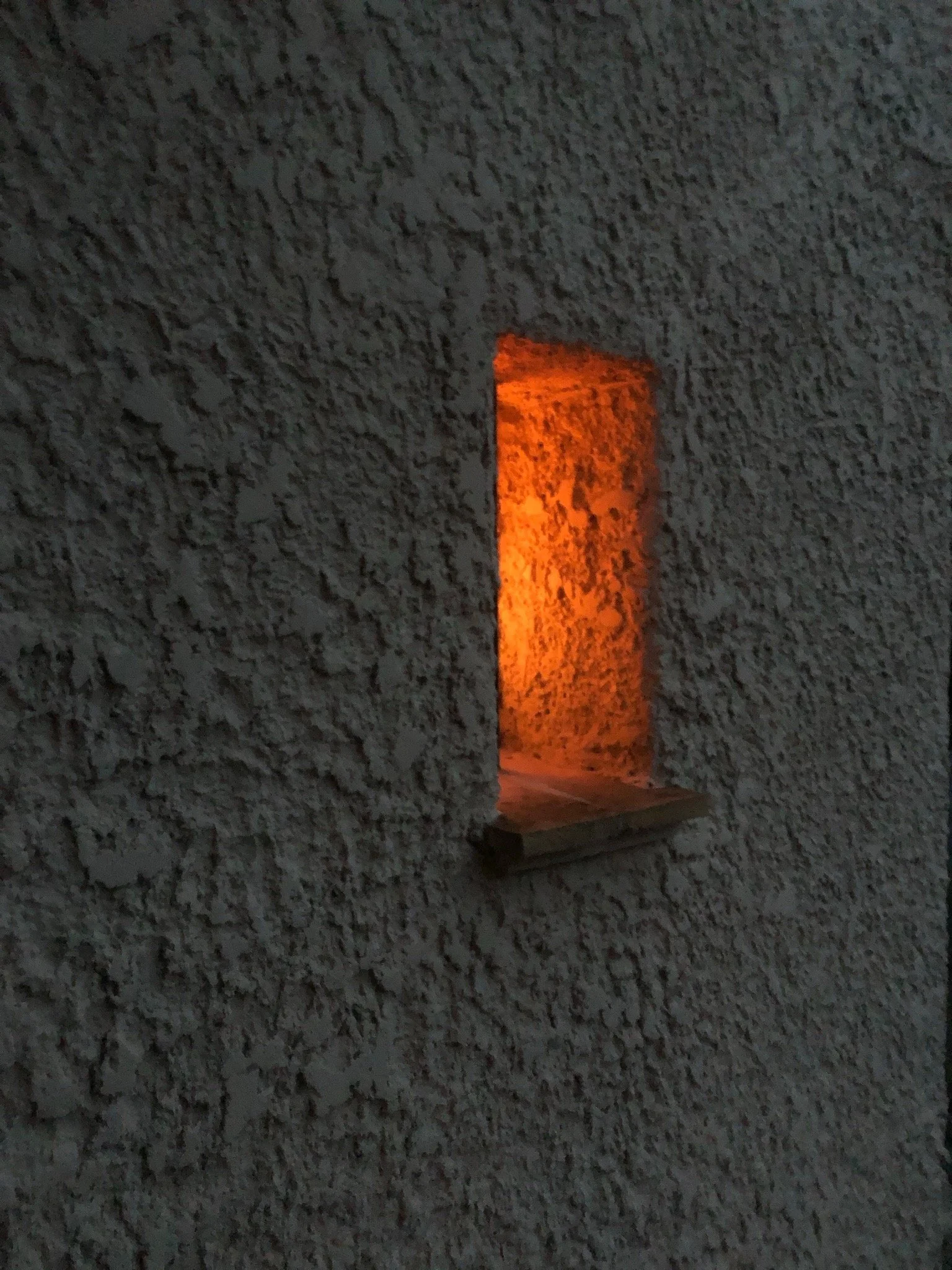 A small rectangular opening in a rough-textured wall with an orange light emanating from inside.