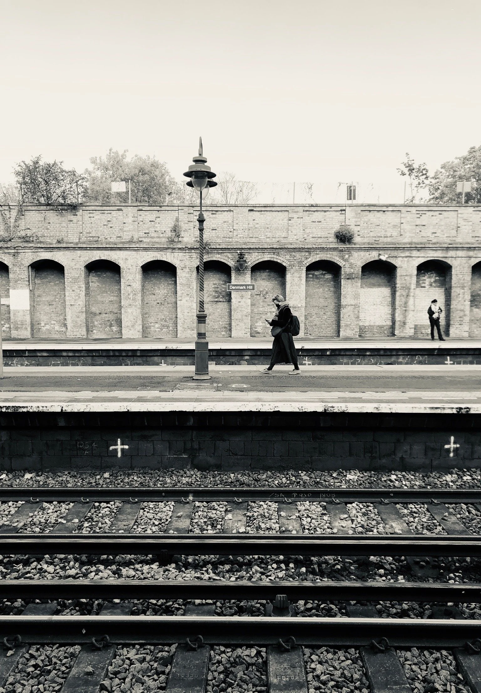 Black and white photo of a train station platform with a woman walking and looking at her phone, another person standing in the background, and a brick wall with arched openings. Railroad tracks are in the foreground.