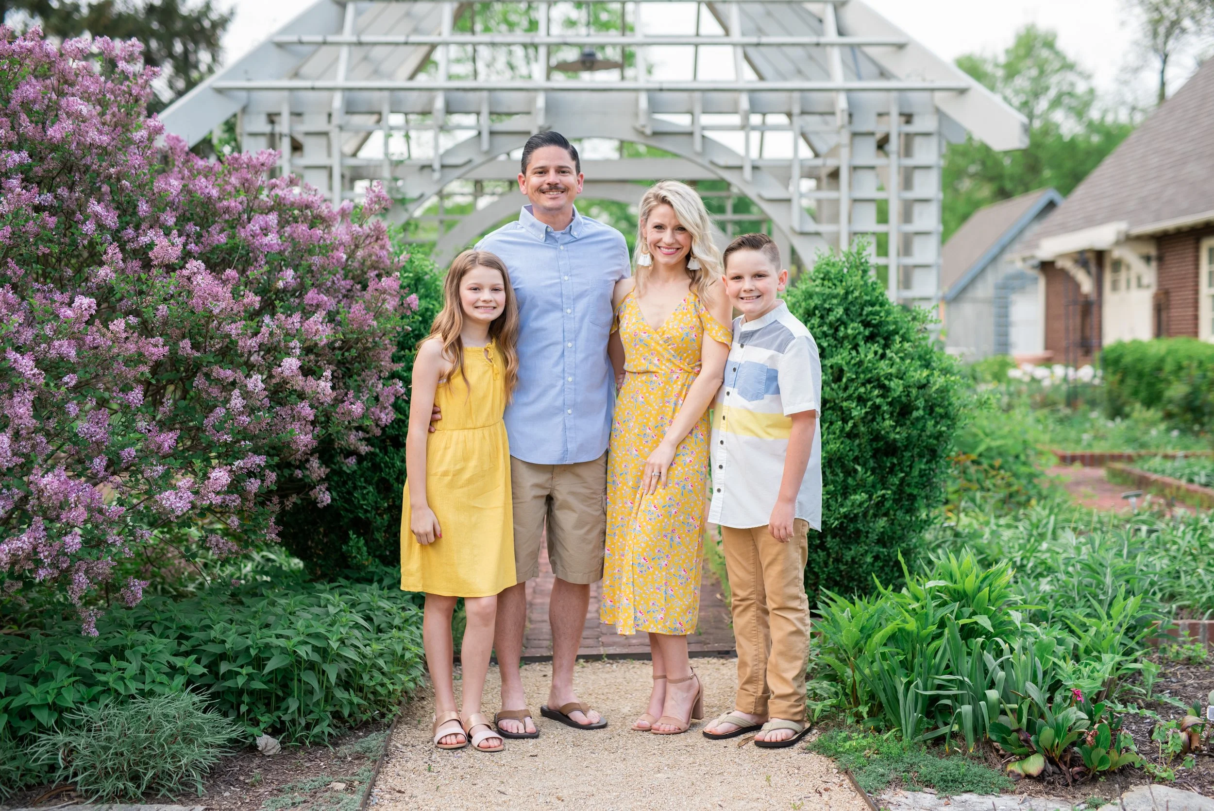 A family of four standing on a garden path surrounded by blooming flowers and greenery, under a white arbor in a backyard.