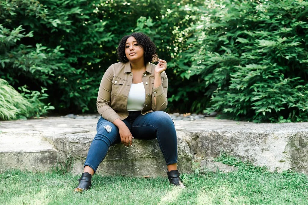 A woman sitting on a stone in a lush green outdoor setting.