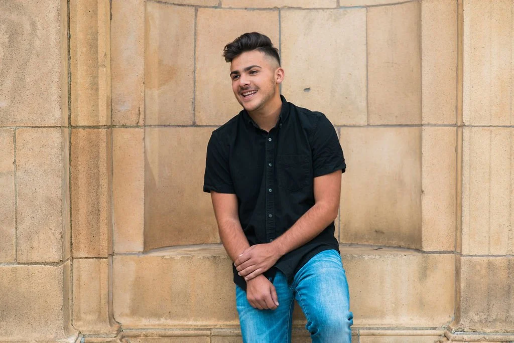 A young man with short dark hair and a beard is smiling while leaning against a beige stone wall. He is wearing a black short-sleeved button-up shirt and light blue jeans.