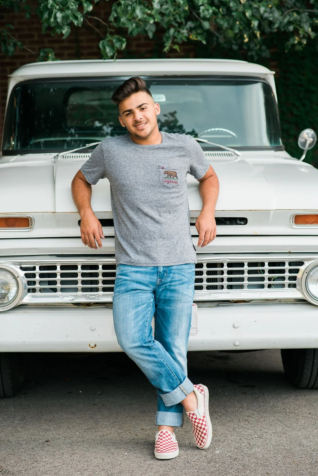 A young man with dark hair, wearing a gray t-shirt with a California bear graphic and jeans, is leaning against the front of a vintage white truck, smiling at the camera.