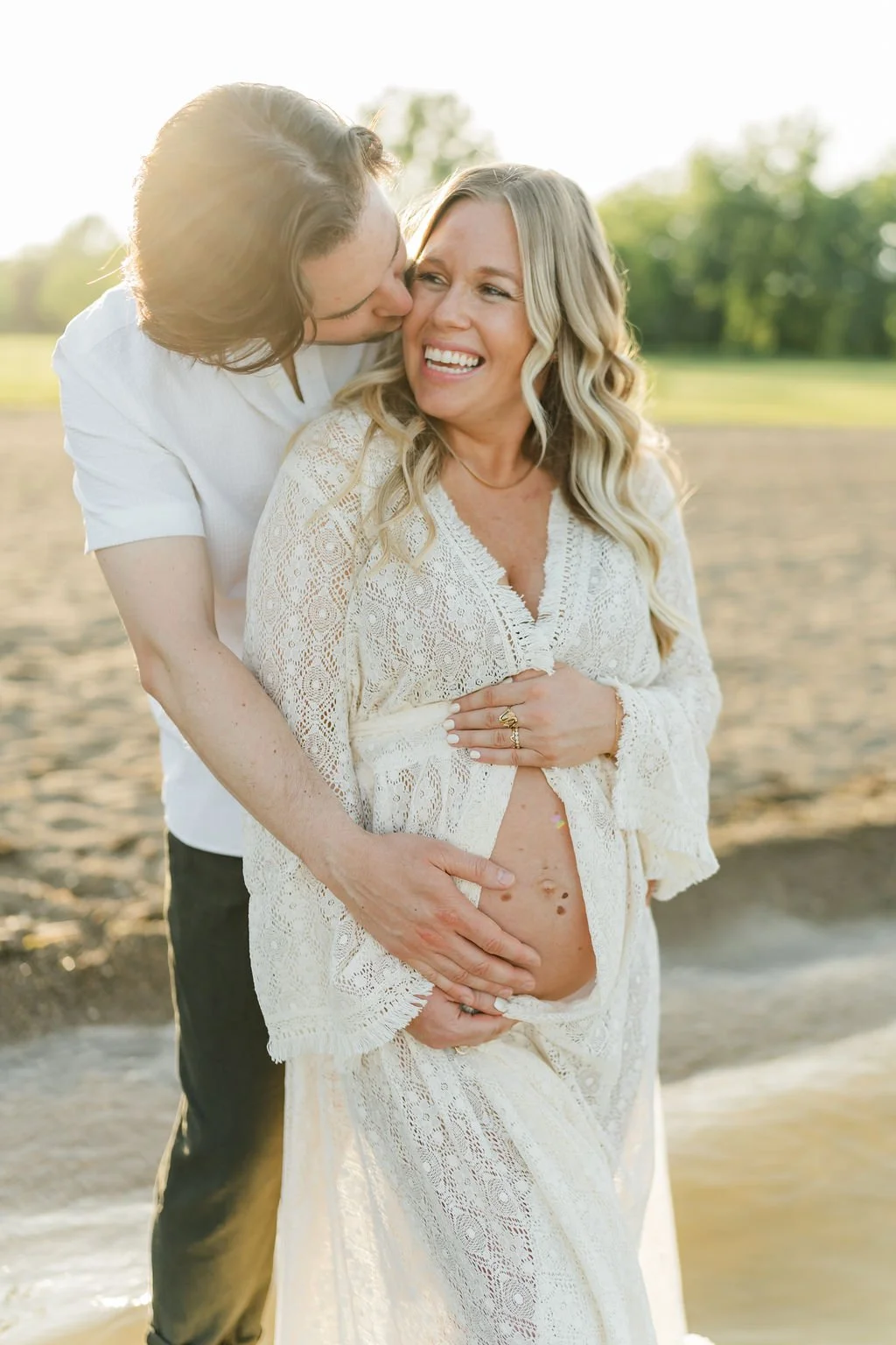 Pregnant woman in a white lace dress smiling with a man behind her kissing her cheek at sunset outdoors.