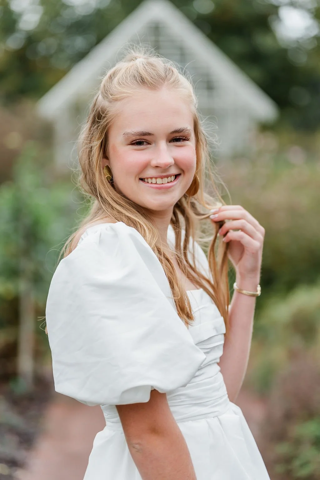 A young woman in a white dress smiling outdoors with a blurred green background.