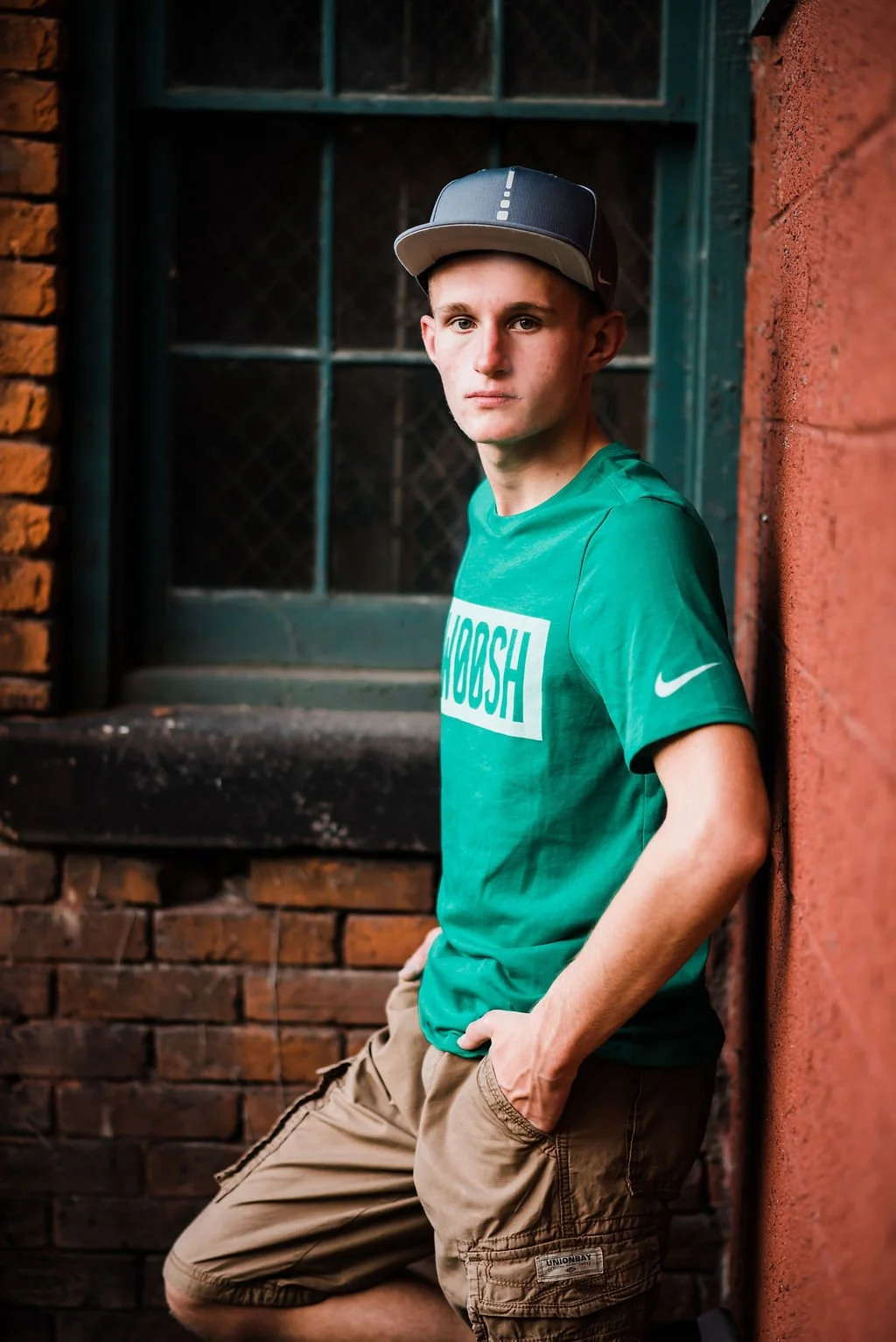 A young man with short hair, wearing a gray baseball cap, teal t-shirt with the word 'NOOSH' on it, and khaki shorts, stands leaning against a red brick wall near a window with a green frame and grilles. The background includes a brick building exter