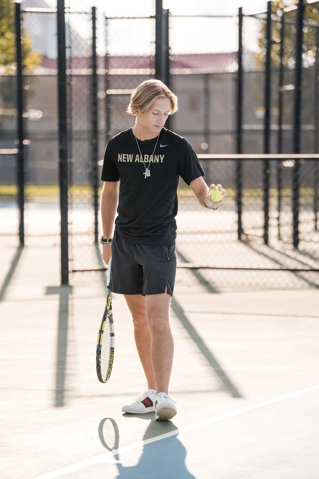 A young man with shoulder-length blond hair on a tennis court, holding a tennis racket in his right hand and a tennis ball in his left hand, dressed in a black T-shirt with "New Albany" printed on it, black shorts, and white sneakers, with a chain ne