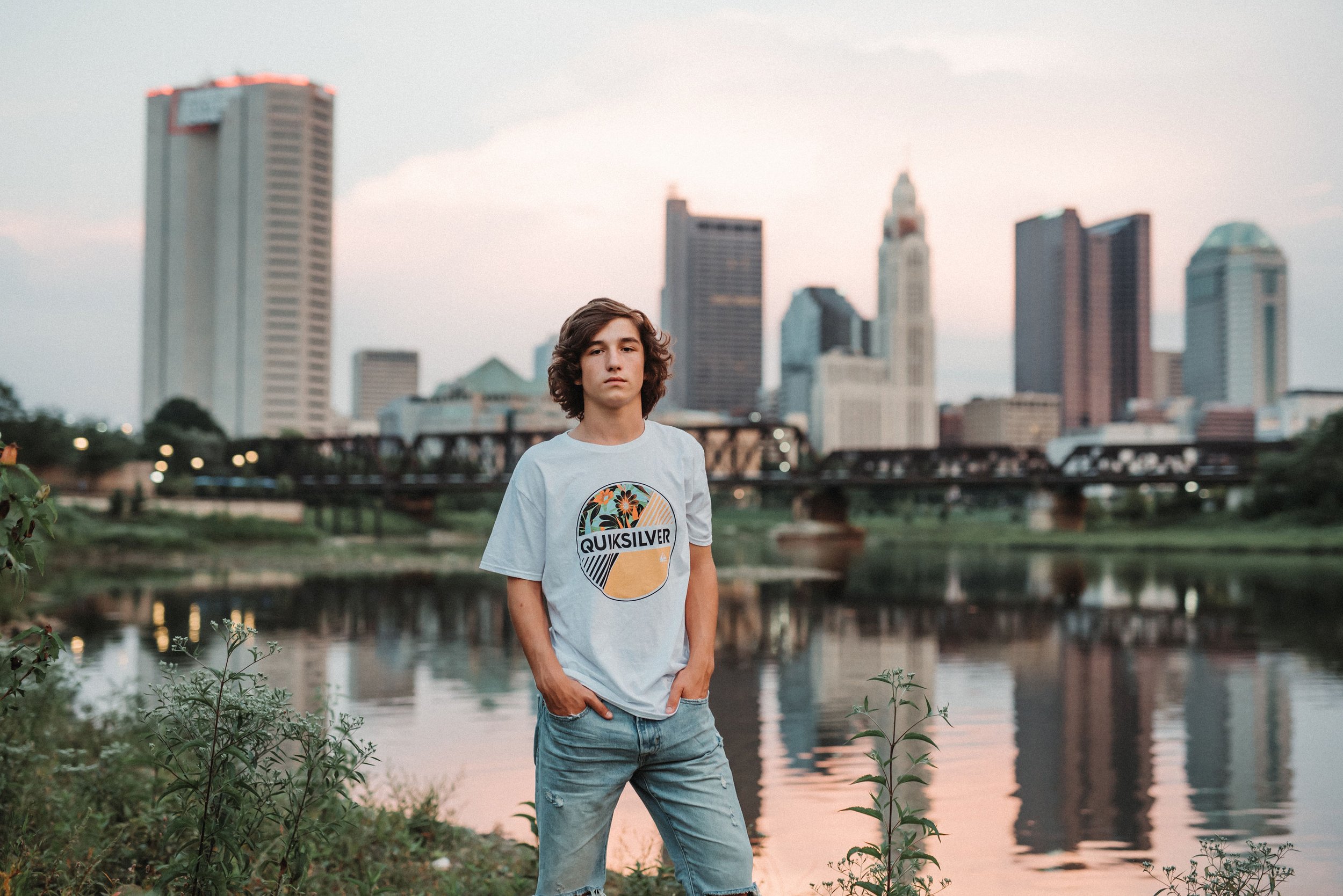 A young man standing by a river with a city skyline in the background during sunset.