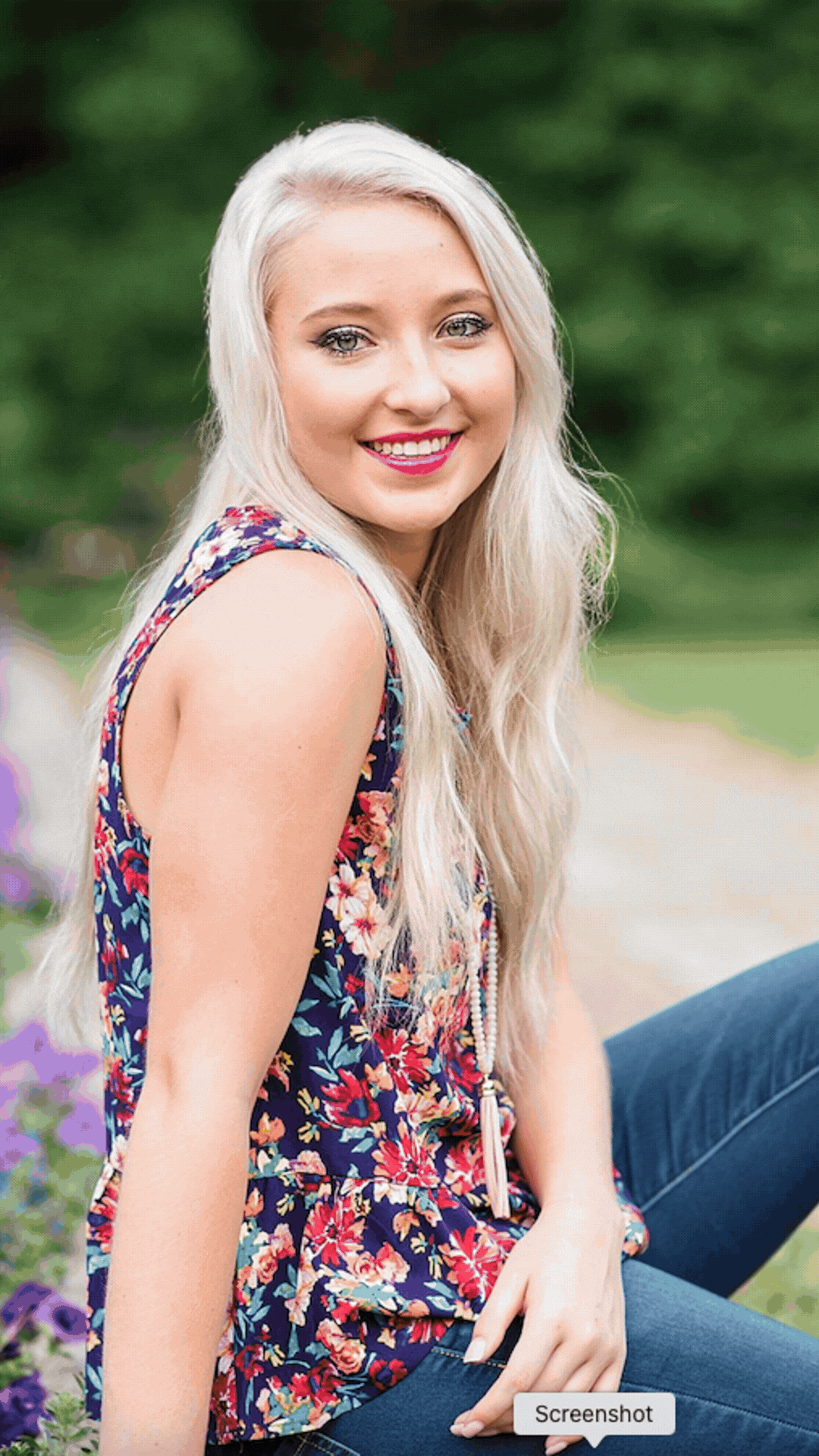 A young woman with long, platinum blonde hair sitting outdoors, smiling, wearing a sleeveless floral top and blue jeans, with a green blurred background.
