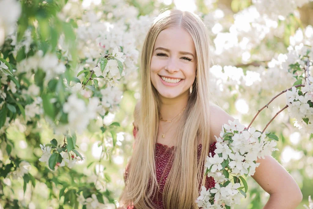 A young woman with long blonde hair smiling outdoors among white flowering bushes.